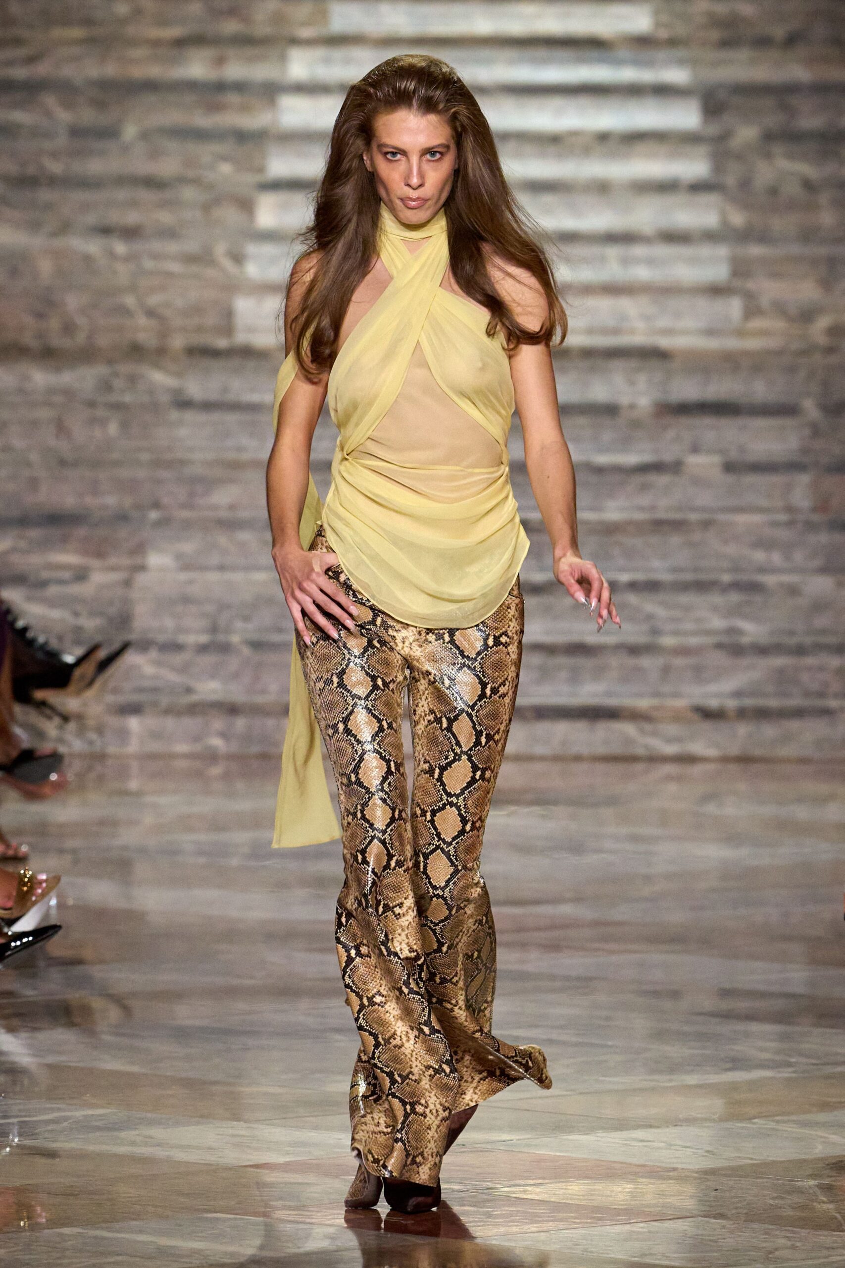 Butter-yellow chiffon halter top, crossed bands over a sheer nude panel. Brown python-print trousers, cut long enough to puddle over the foot. Pointed dark boots just breaking the hem. Walking into a hotel bar while the marble floor still holds the afternoon heat.