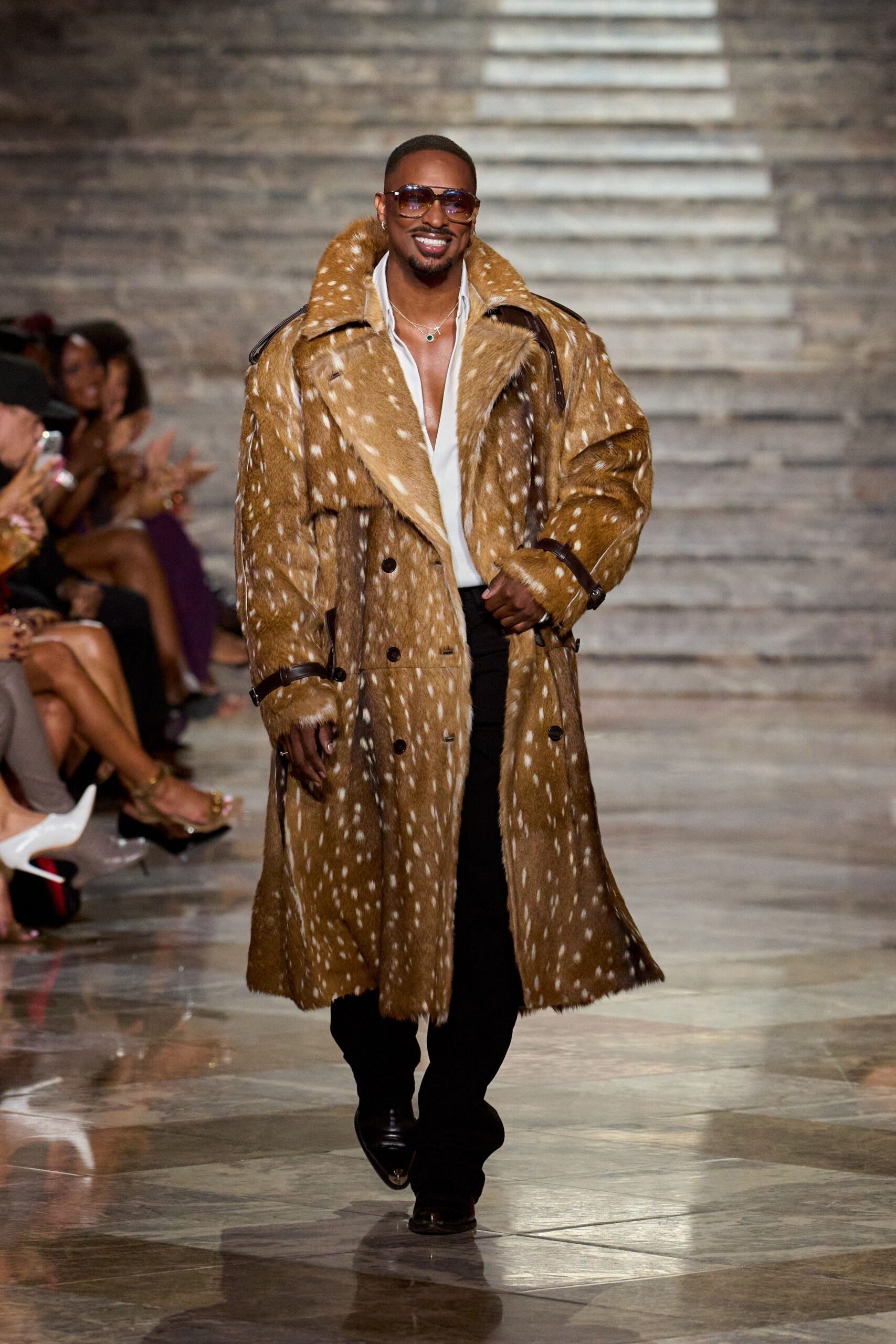 Amber gradient aviators with a thick gold bridge. Double-breasted fawn coat in spotted hide, dark leather straps at the cuffs. White shirt worn open deep at the chest. Black flared trousers breaking softly over the shoe. Pointed black boots with a glinting metal toe. For crossing a hotel lobby when everyone looks up at once.