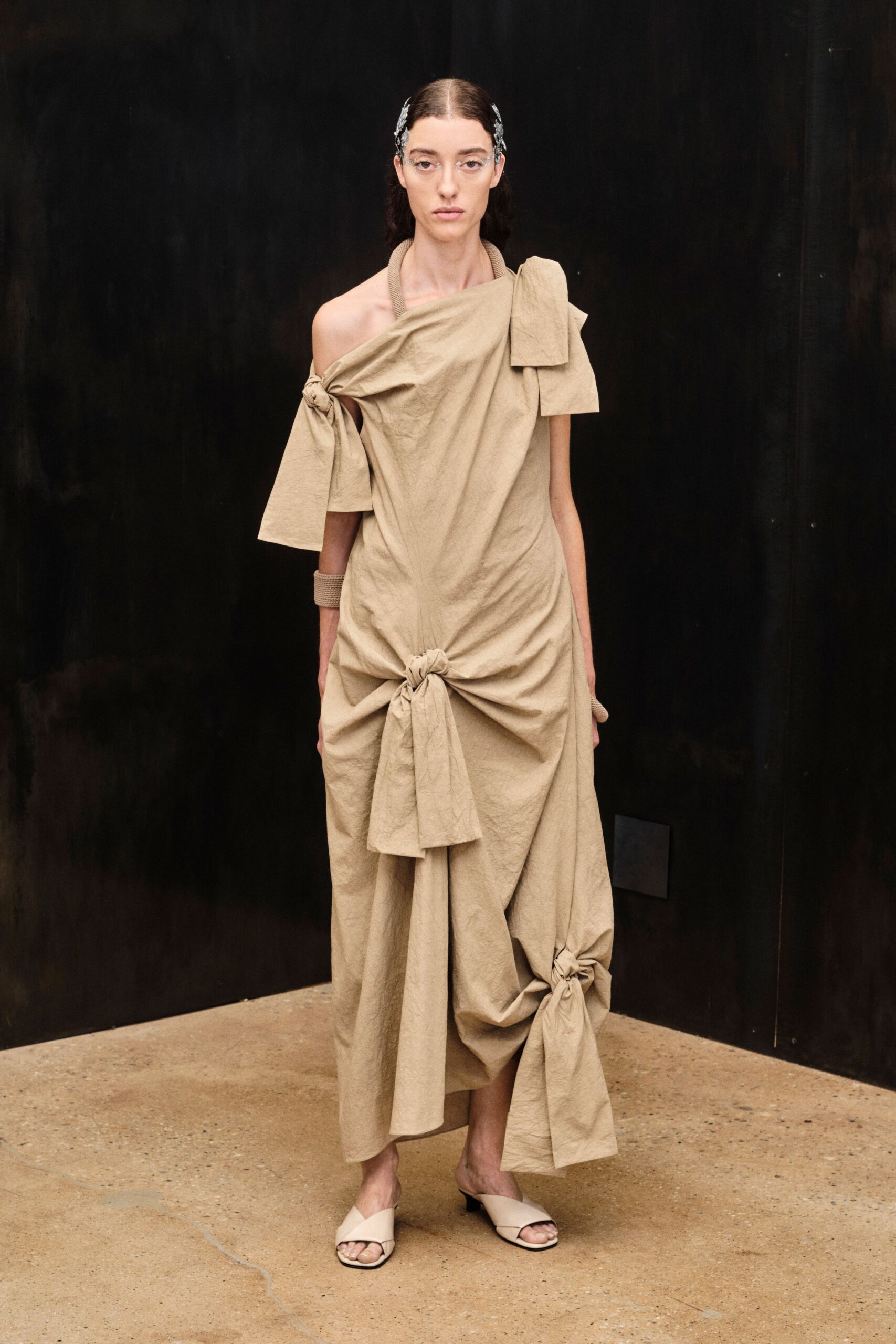 Clear frameless glasses with silver filigree climbing the temples. Sand crinkled dress draped off one shoulder, knotted at hip and hem. Wide ribbed cuff at one wrist. Cream cross-strap mules on a low black kitten heel. Walking across a museum courtyard before the stone has cooled.