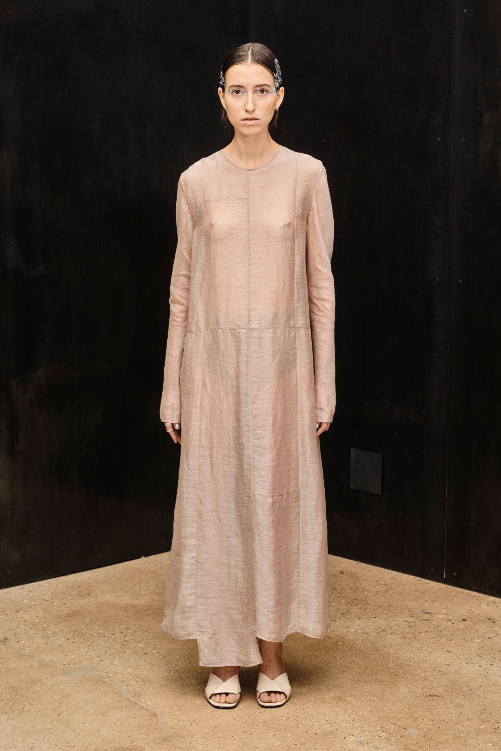 Rimless glasses with silver filigree curling past the outer corners. Blush silk maxi dress, sheer through the bodice with a dropped waist seam. Cream leather mules with blunt folded vamps. Museum atrium hush, shoes clicking louder than anyone's voice.