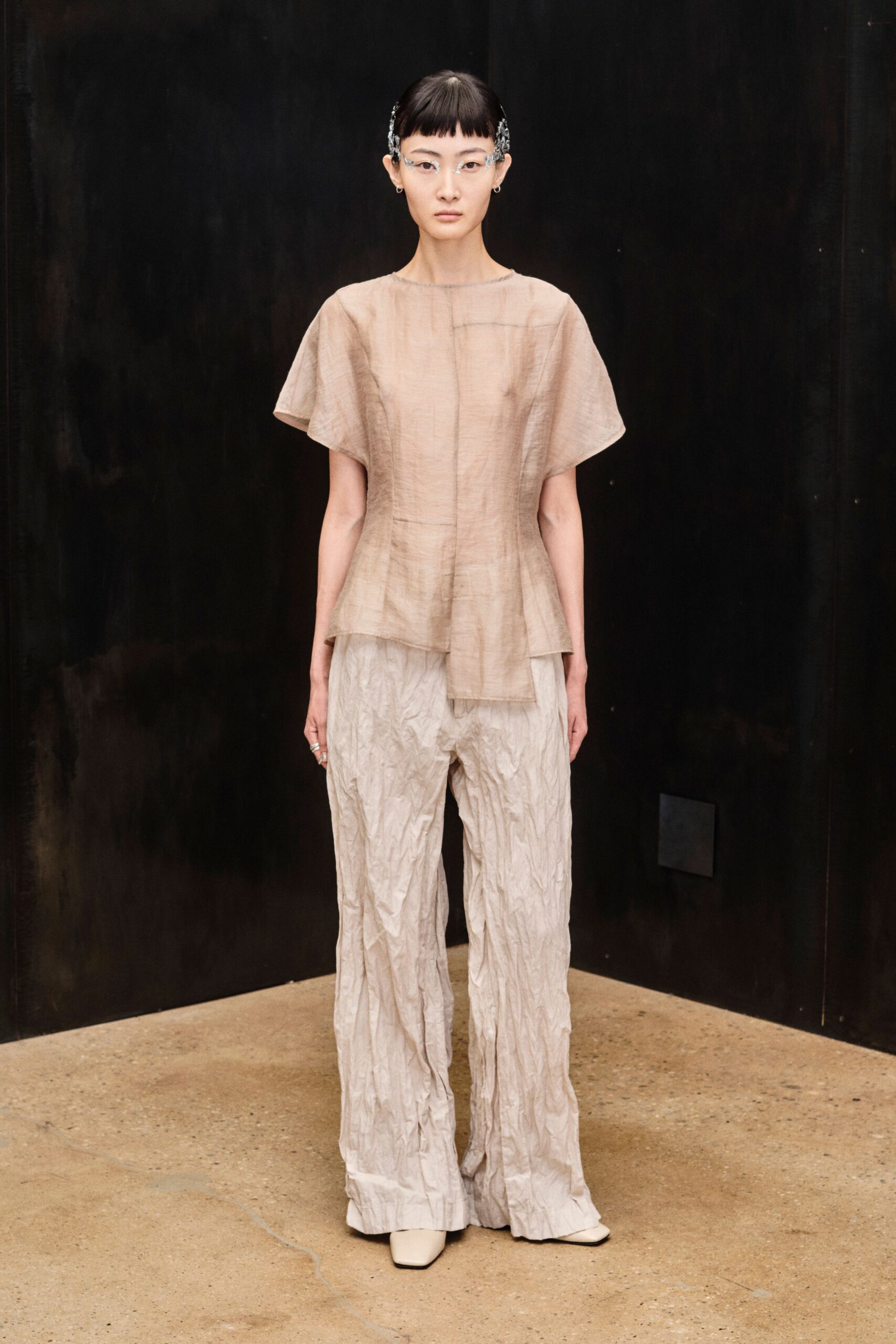 Clear rimless glasses with silver floral sprays at the temples. Sheer taupe top with dolman sleeves and a longer front panel. Pale crinkled trousers, permanently crushed from hip to hem. Square-toe cream boots peeking from the pooled cuff. Quiet hour in a conservatory after the watering cans are put away.
