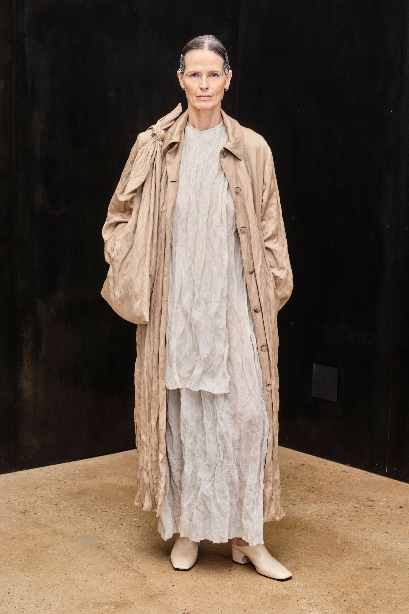 Long parchment coat in crushed silk, a knotted panel slung off one shoulder. Chalk crinkled tunic with a raw, wavering neckline. Matching crinkled skirt falling in soft vertical creases. Cream square-toe ankle boots with low cutout heels. Early museum opening, concrete still cool from the night.