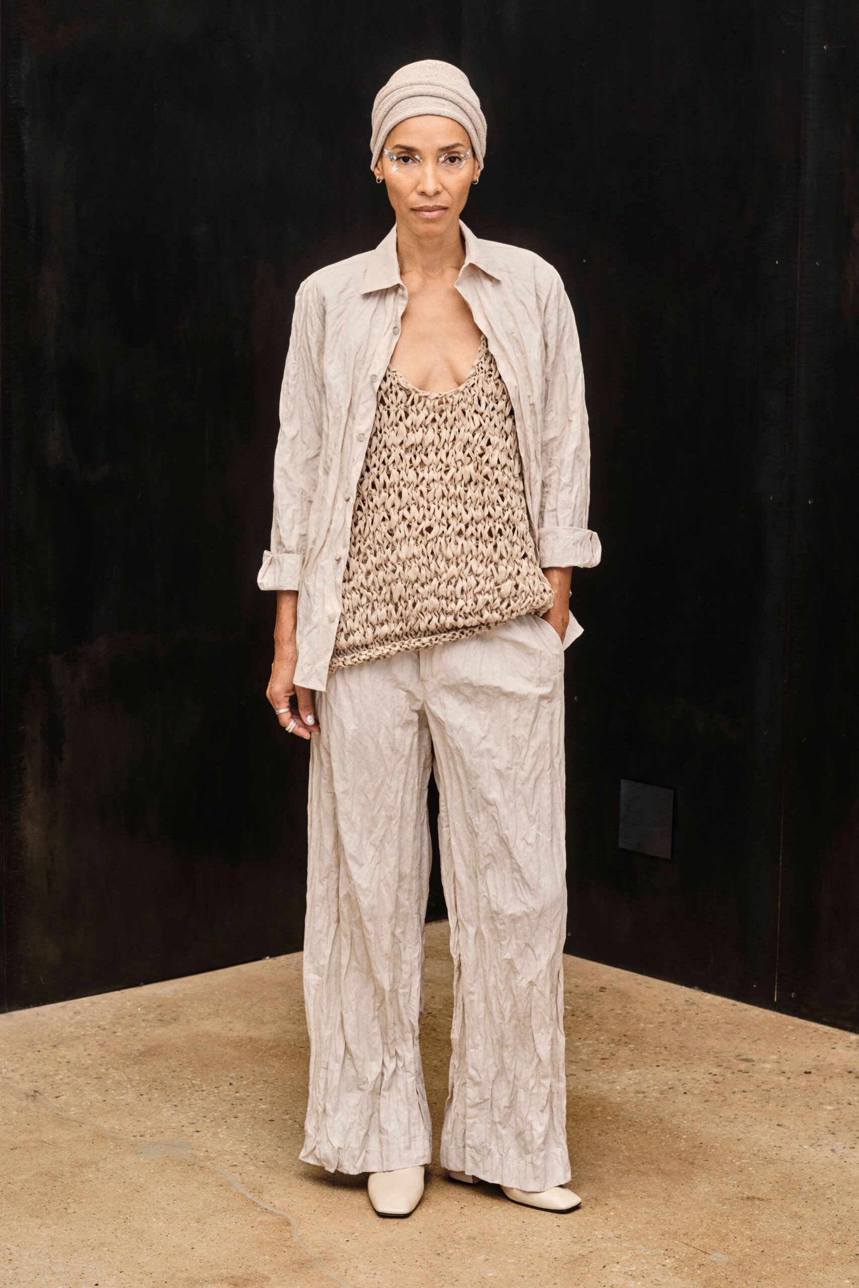 Taupe jersey headwrap layered in soft horizontal folds. Pale crinkled shirt worn open, sleeves rolled into blunt cuffs. Loose open-knit tank in chunky ribbon yarn, hem curling under. Matching wide trousers in the same permanently creased cloth. Cream square-toe boots. For the first cool morning after a week at the coast.