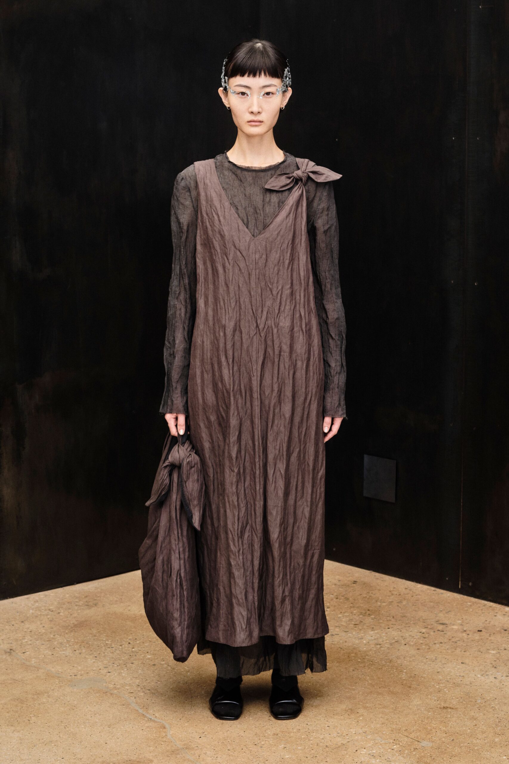 Clear frameless glasses with crystalline side sprays at the temples. Mauve crinkled slip dress, a bow knotted on one shoulder. Smoke sheer long-sleeve underlayer with a softly rolled raw neck. Oversized matching sack, gathered into another bow. Black square-toe flats. Walking through a museum atrium just before the rain starts.