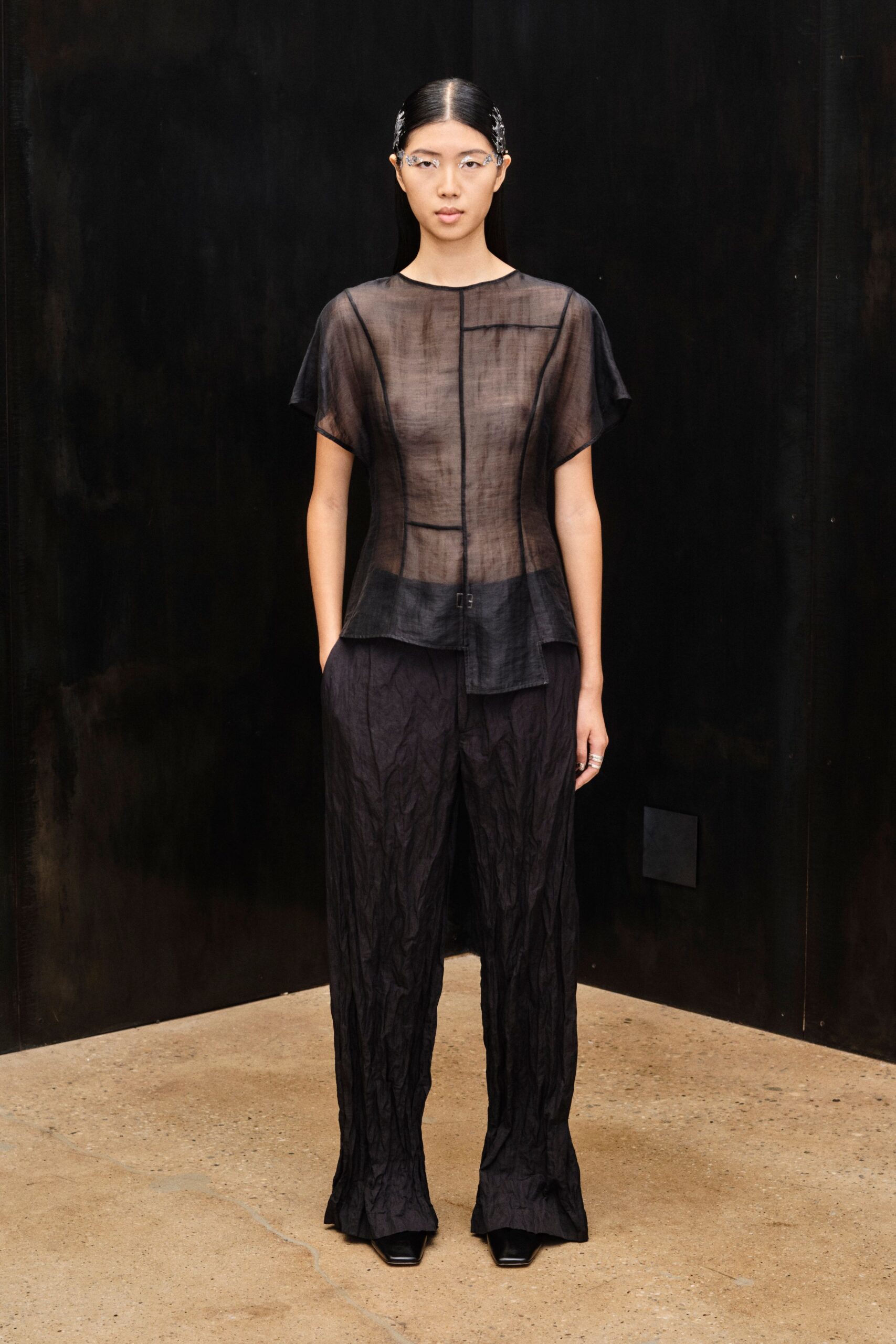 Clear rimless glasses with silver scrollwork curling off the temples. Smoke-black gauze top, sheer and paneled with narrow binding lines. Crinkled black trousers, pooling lightly over the square toes. Black square-toe shoes. Walking through a museum after closing, hearing your steps on concrete.