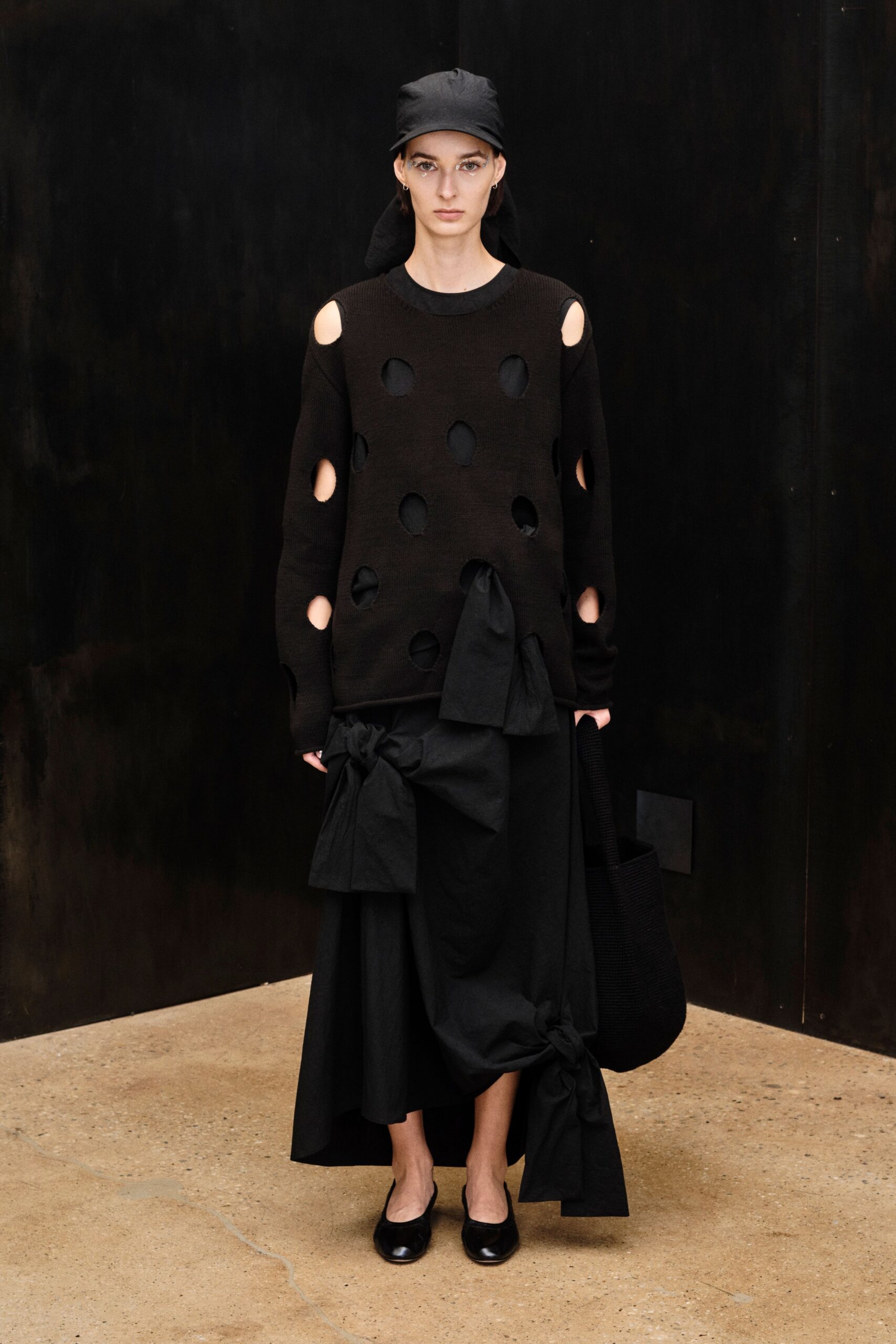 Black cap with a long draped back panel. Black rib knit sweater punched with raw circular cutouts. Full black skirt in crisp cloth, hitched into oversized knots. Deep black woven tote tucked under one arm. Square-toe black flats. For crossing a sculpture court before the rain starts.