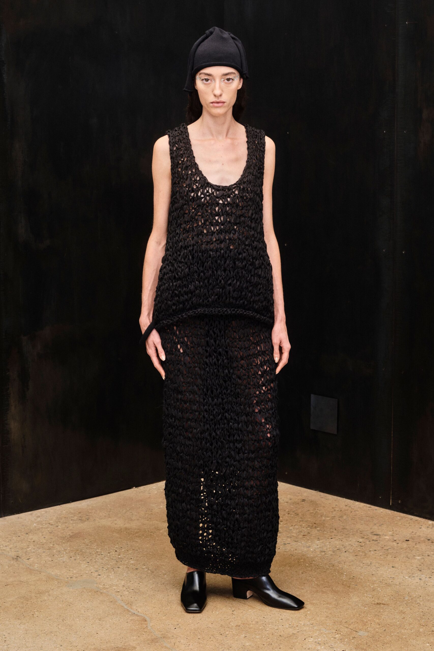 Black knit cap with ear-like flaps hanging beside the temples. Clear wraparound frames. Loose black open-knit tank, scooped low and curling at the hem. Matching open-knit column skirt, sheer through the legs. Black square-toe ankle boots with a low block heel. Crossing a concrete courtyard after dark, still damp from the hose.