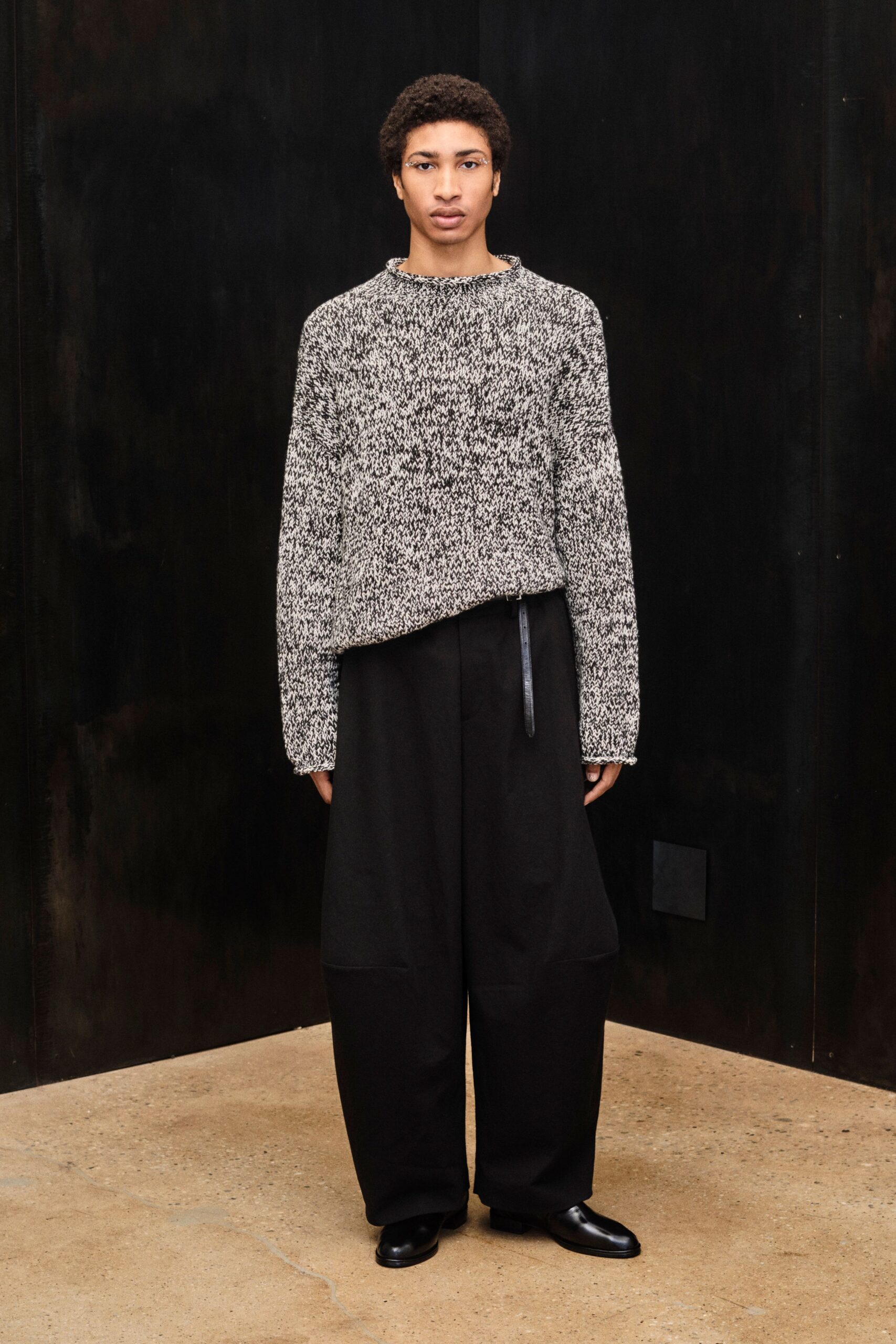 Black-and-cream marl knit sweater with rolled edges and an uneven hem. Black full trousers with rounded outseams and a slim leather tab hanging front. Square-toe black leather slip-ons, low and blunt. Walking across a concrete lobby before the heating has kicked in.