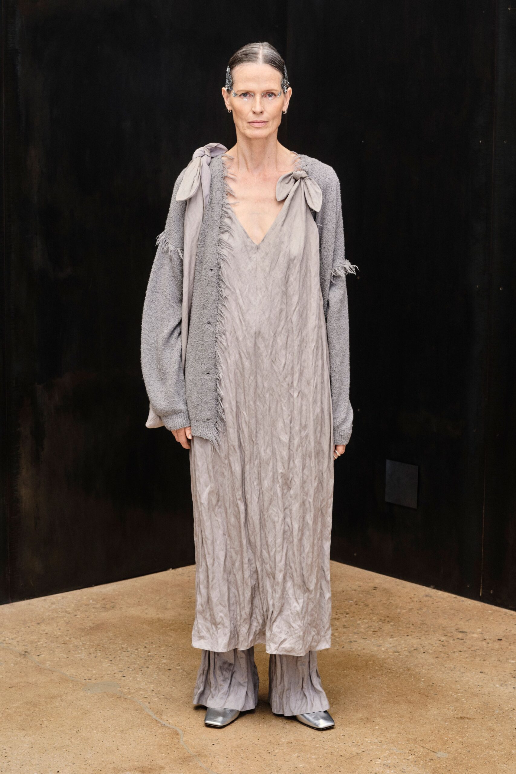 Pebbled dove-grey knit cardigan, seams and sleeve slits left feathery. Crinkled ash slip dress with bow-tied shoulders and a frayed V. Matching crushed trousers pooling under the hem. Square silver boots flashing from beneath. For crossing a museum courtyard just after the rain stopped.