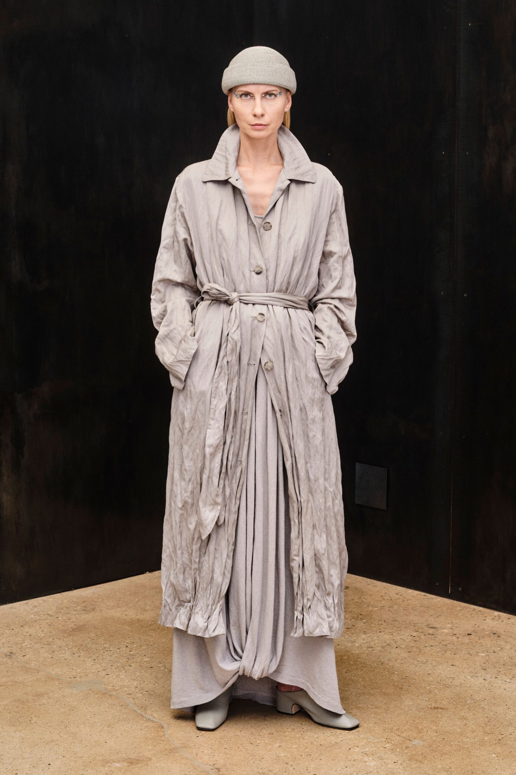 Pale ribbed knit beanie rolled thick above the brow. Ash crinkled coat with a high spread collar and self-tie waist. Long dove dress falling in heavy folds, one twisted panel pooling front-center. Square-toe ash leather boots with a low block heel. Crossing a quiet museum courtyard just after the rain stopped.