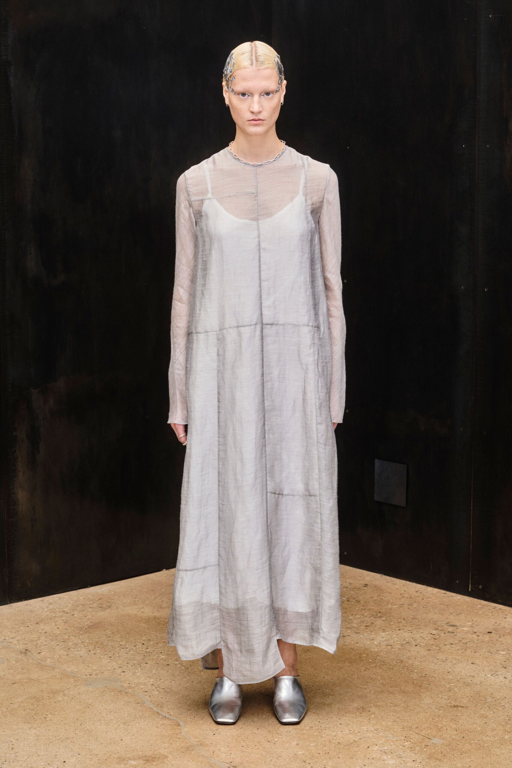 Fine silver chain sitting close at the collarbone. Sheer ash-grey gauze dress, center seam dropped into a low yoke. Chalk slip showing softly through the translucent bodice. Square-toe silver flats with a liquid-metal shine. Early ferry crossing, wind flattening everything against the body.