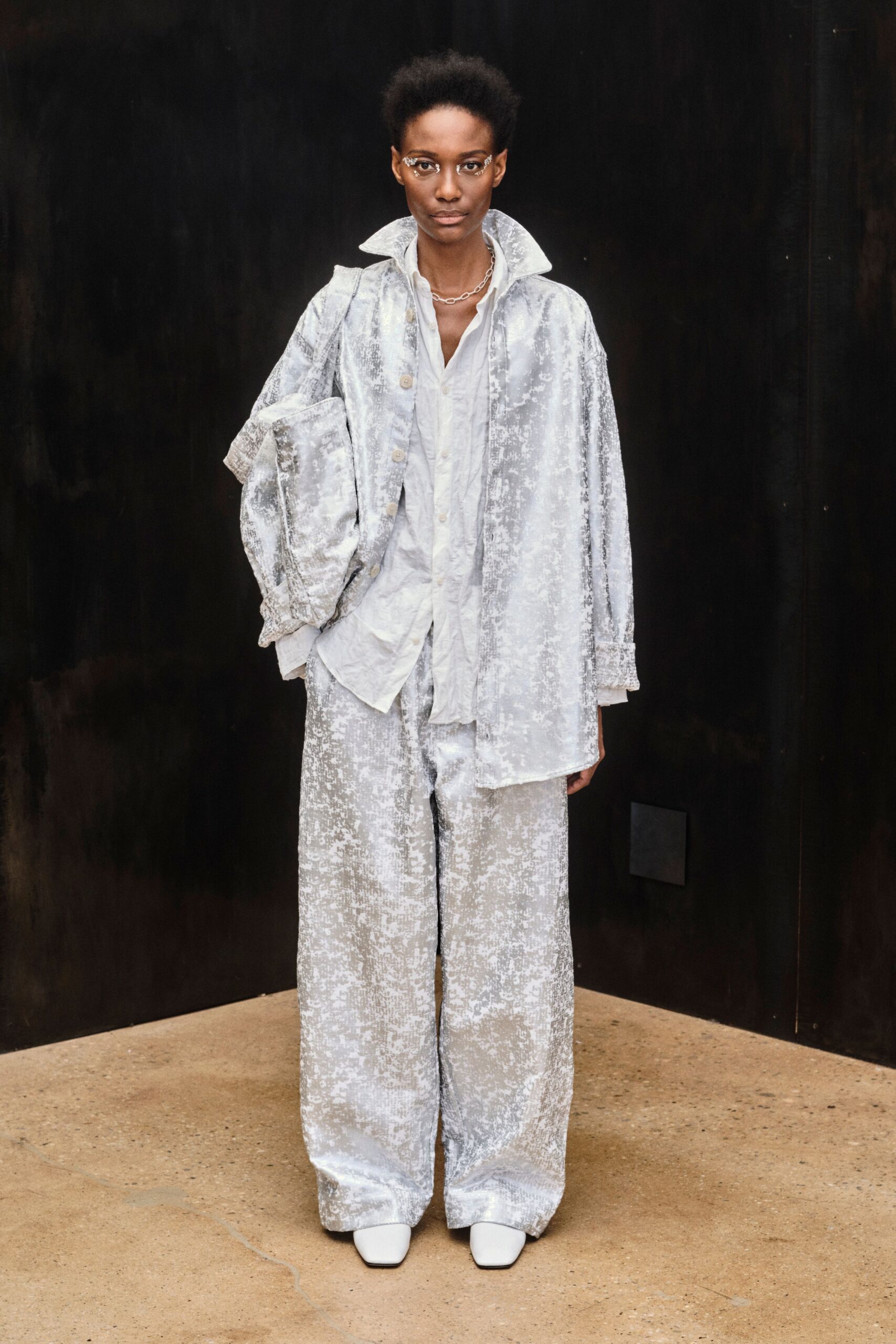 Powdered silver crushed velvet coat, collar kicked high and cuffs turned back. Soft white shirt, rumpled placket loosening into an open V. Matching wide-leg velvet trousers, pooling heavily over the shoes. Short oval-link silver chain. White square-toe boots. Cold morning in a concrete atrium before the building fully wakes.