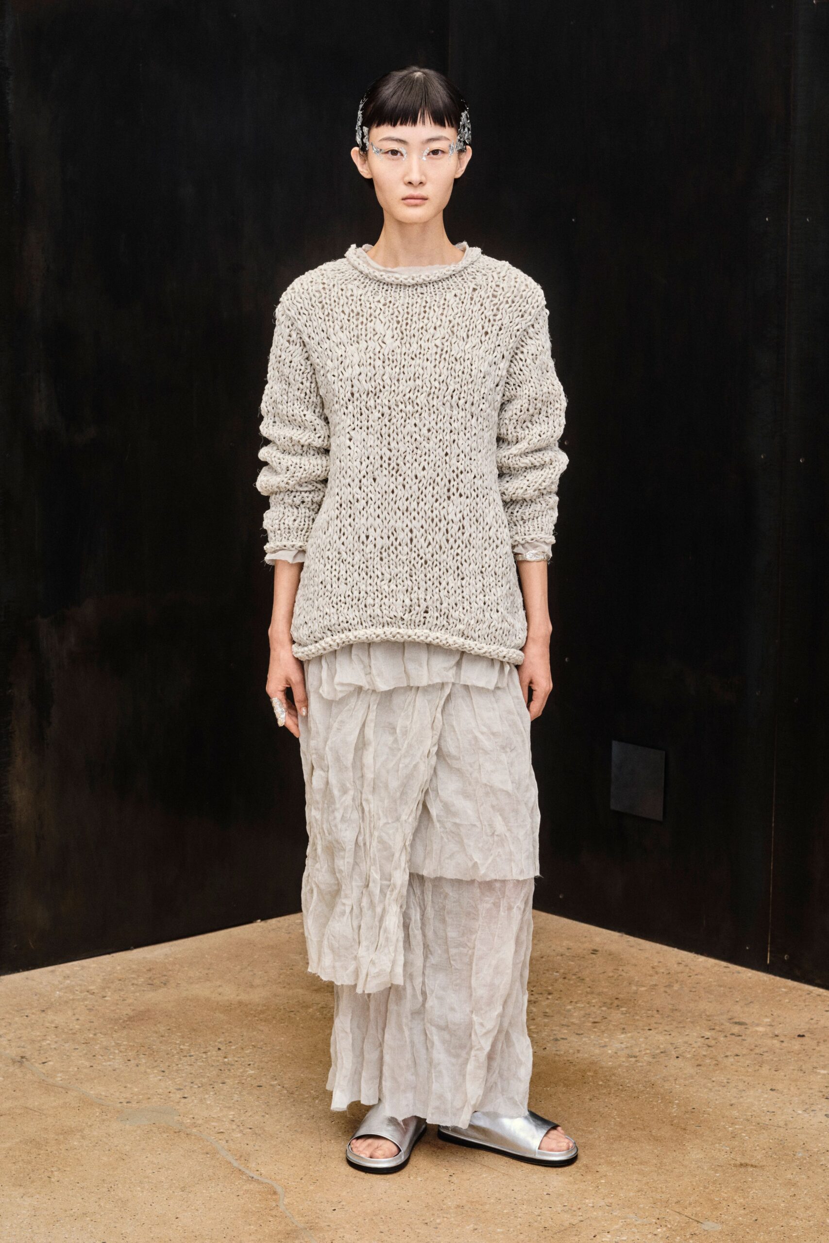 Rimless glasses hooked with silver foil-like ear pieces. Oatmeal open-knit sweater, collar rolled into a soft rope edge. Cloud-white crinkled skirt layered in uneven translucent tiers. Silver leather slide sandals. Crumpled silver ring. Laundry still warm from the line, windows open before the rain.