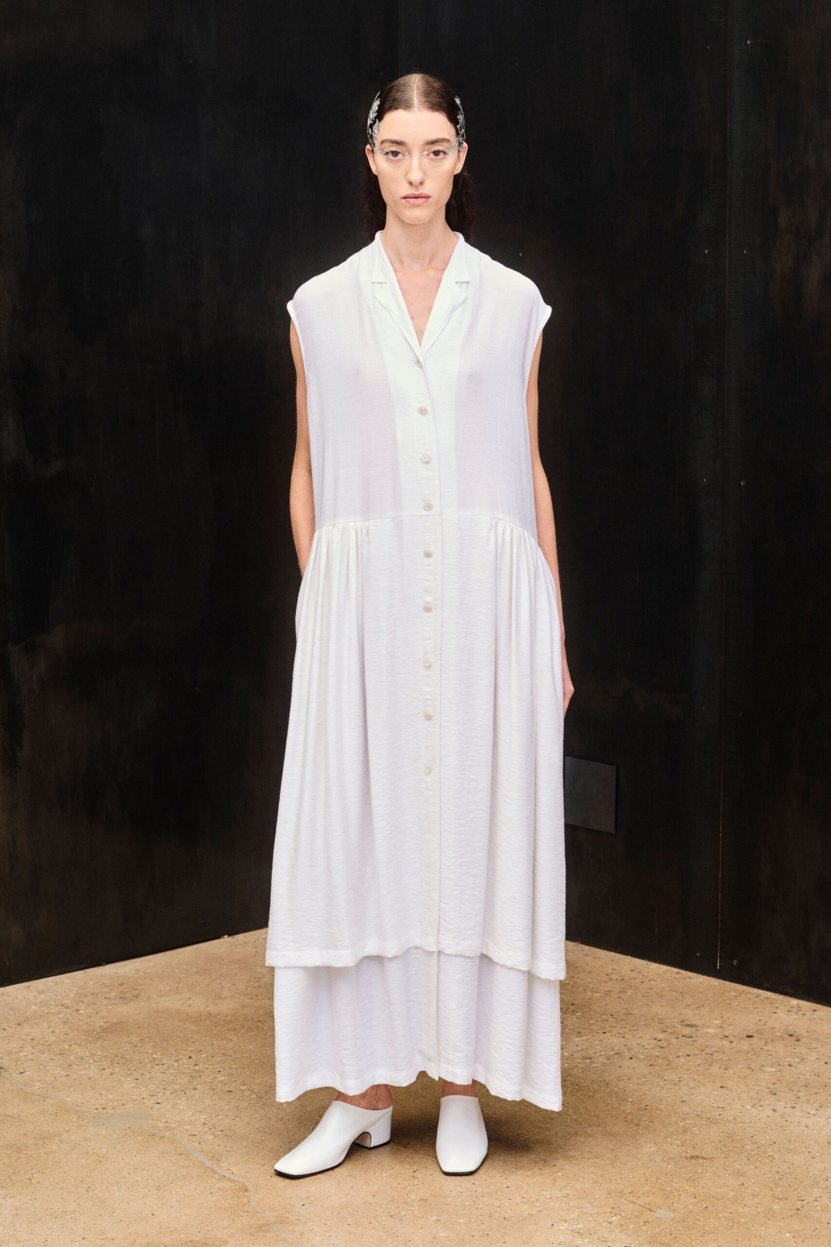 Clear crystal sprigs pinned tight at both temples. Chalk-white sleeveless shirt dress in gauzy crepe, dropped waist softly gathered. Wide white trouser hem peeking below the dress. Square-toe white leather mules on a low block heel. Early check-in at a desert spa, lobby still cool from the night.