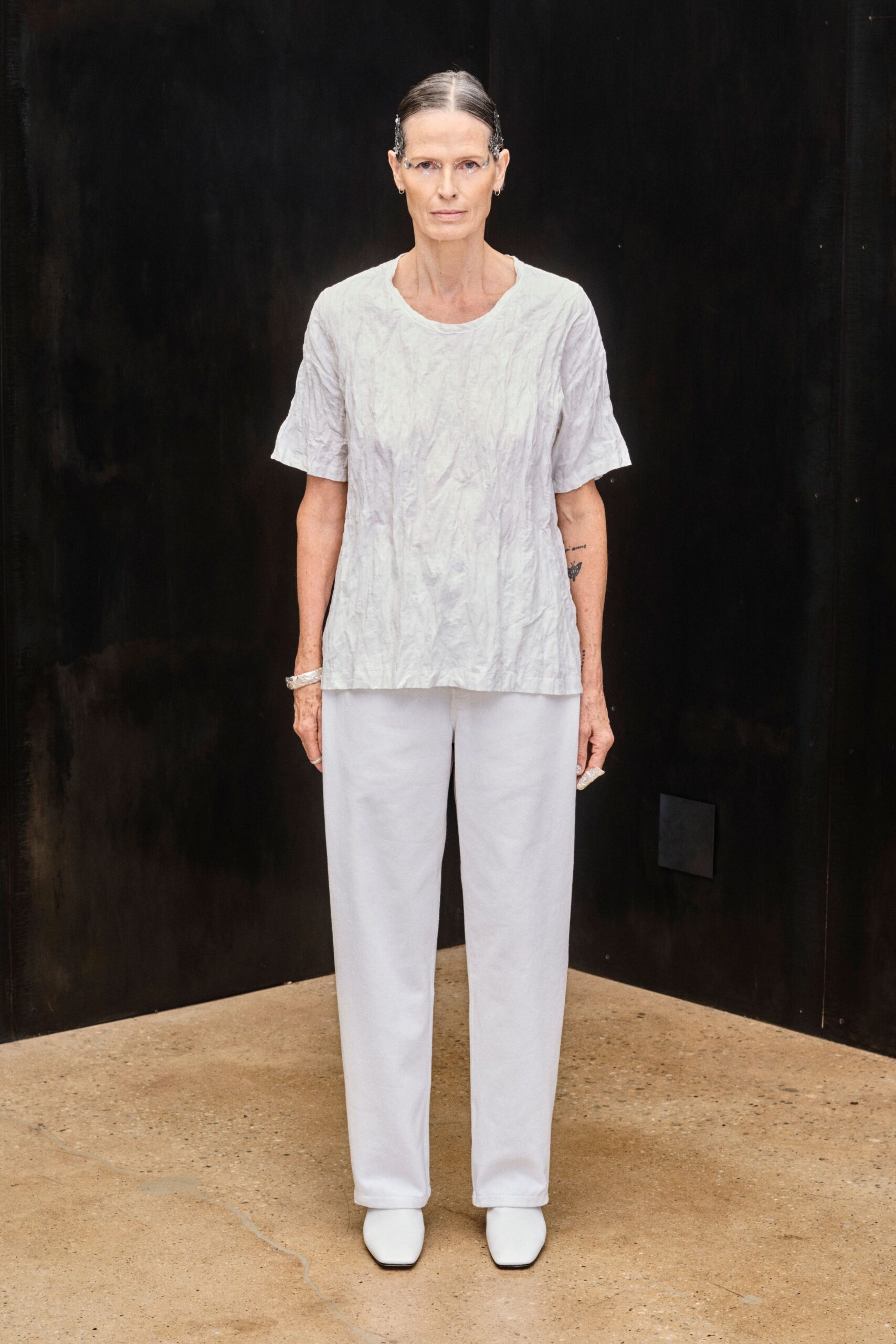 Clouded ice-dye tee, crinkled all over with a softly dropped shoulder. Pale straight trousers, clean front falling almost flush over the shoe. White square-toe boots with a blunt, apron-like front. Morning appointment in a concrete building that still smells faintly of rain.
