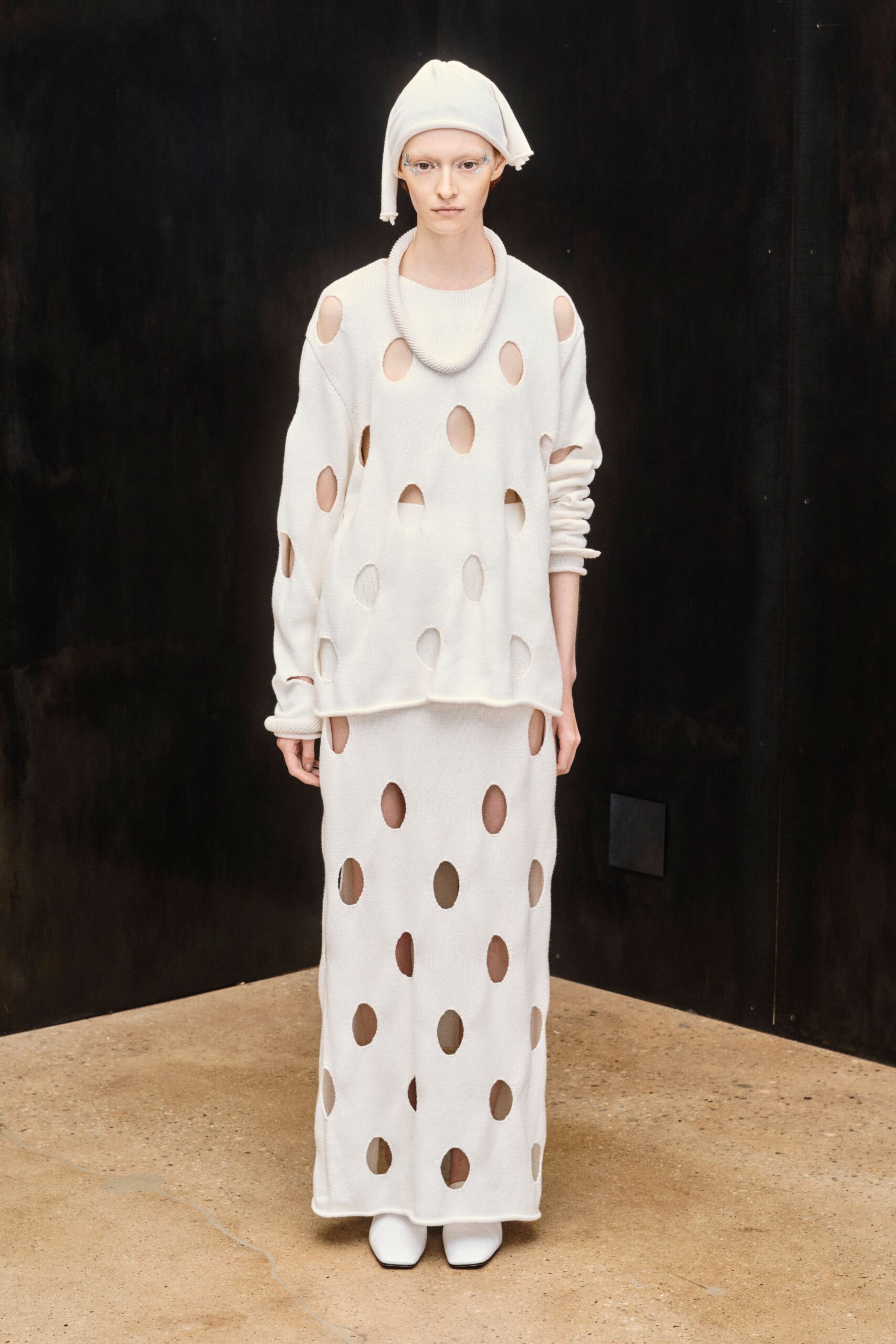 Soft white jersey headwrap, one tail drooping beside the ear. Ivory knit sweater punched with raw-edged oval holes and a rolled neckline. Matching column skirt, the same cutouts marching to the hem. White square-toe boots disappearing under the knit. Museum atrium at opening hour, before the heat comes up.