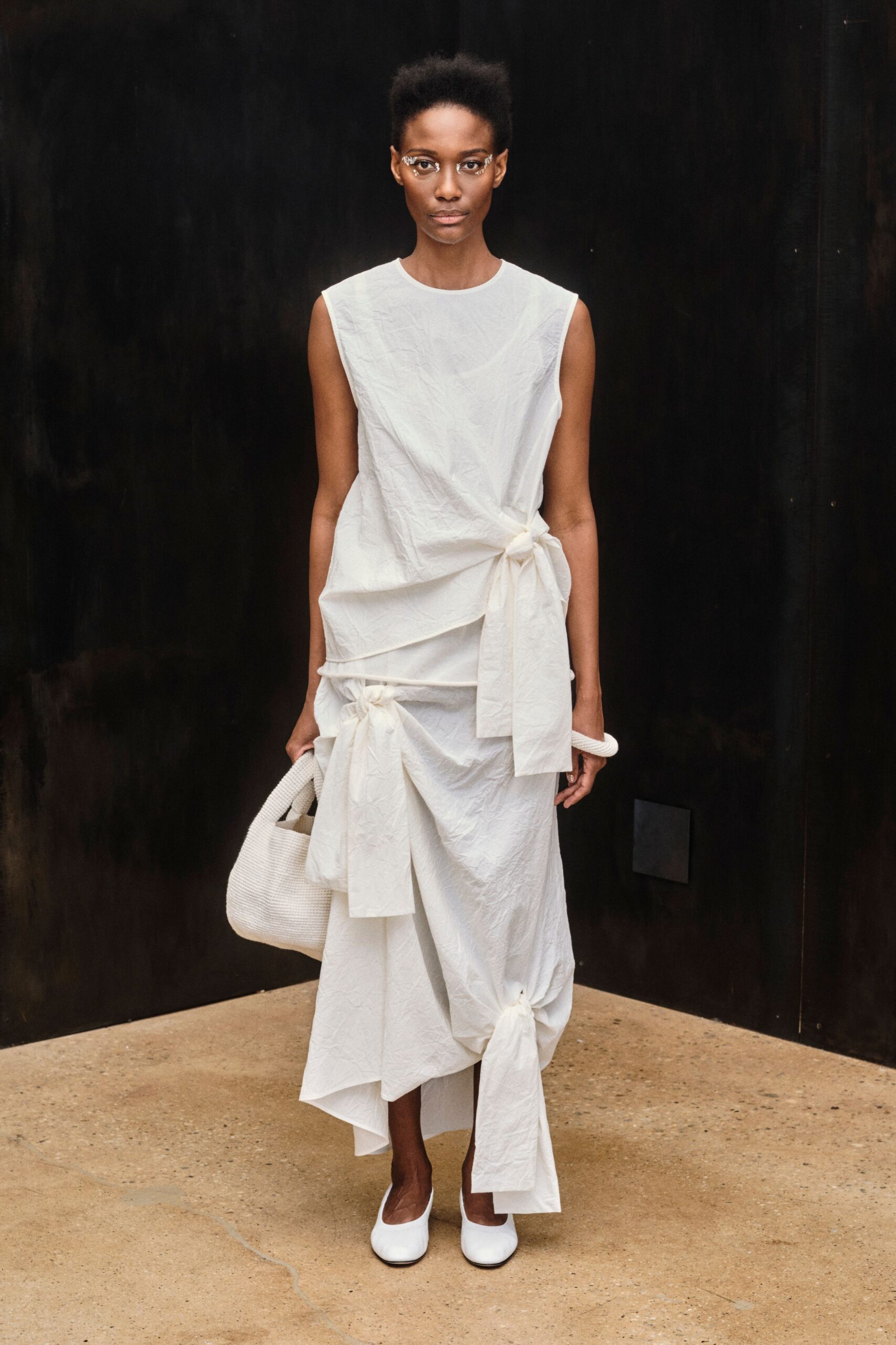 Silver crystal swirls framing the outer corners of both eyes. Crisp white cotton dress, wrinkled and knotted at hip, thigh and hem. Chunky white coil bracelet. Soft white woven tote slung low from one hand. White square-toe flats. Walking across a quiet museum courtyard after the fountain has been switched off.