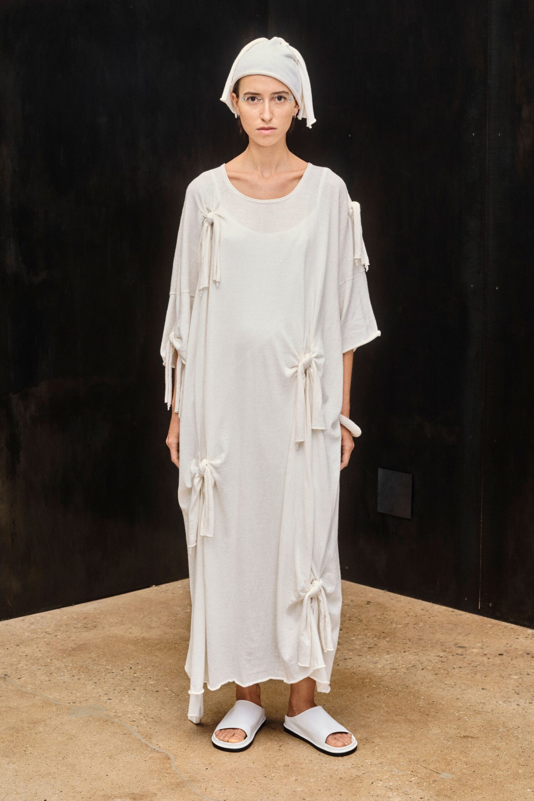 Soft white knit wrapped turban-style, one corner drooping over the temple. Milk-white jersey column dress, gathered into knotted ties down the body. White leather slides with thick molded straps. For carrying a tray of towels through a bathhouse before sunrise.
