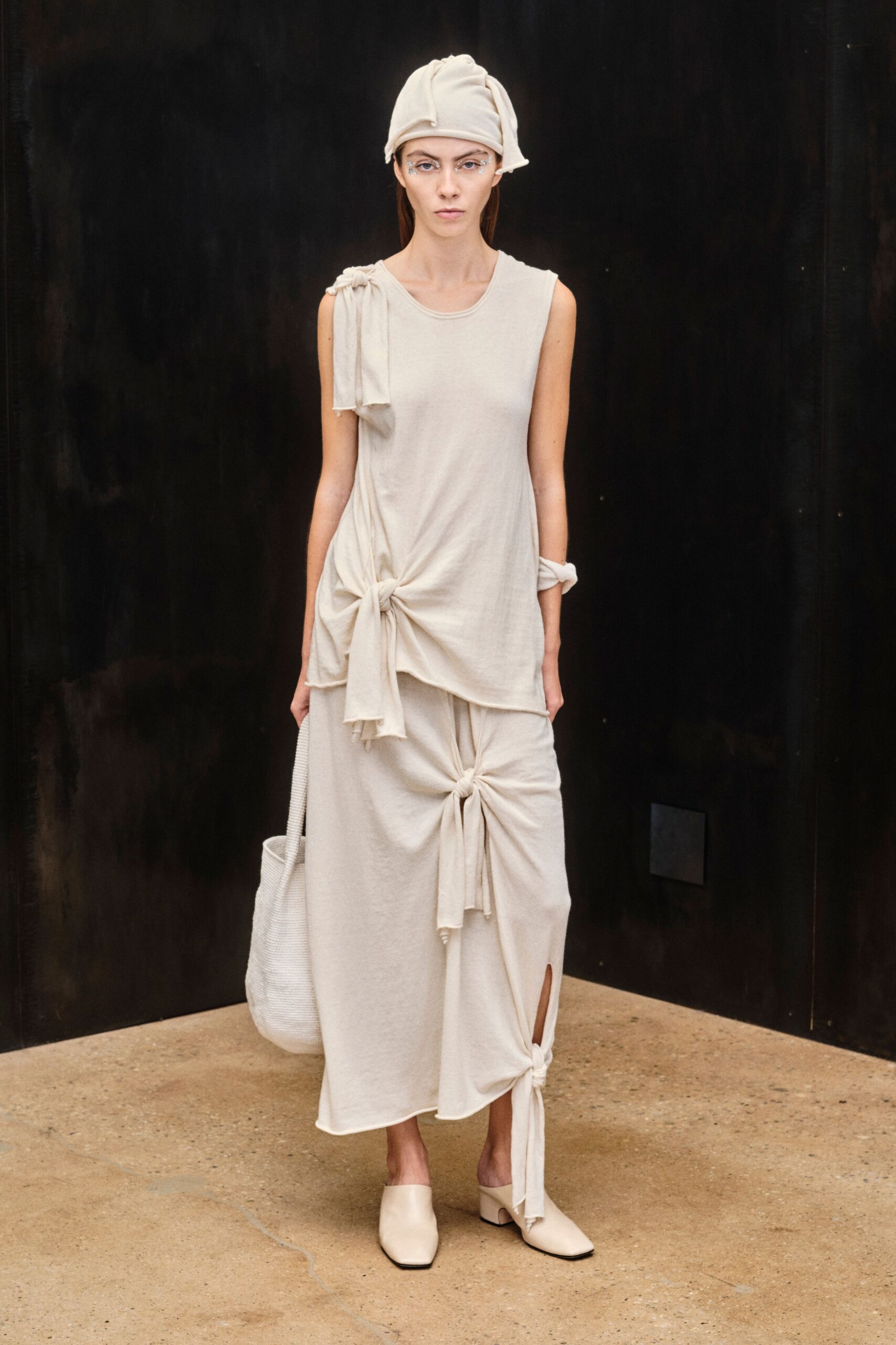 Soft ivory jersey cap, rolled edge kicked up at one side. Sleeveless bone knit dress, knotted at shoulder, hip and shin slit. White ribbed tote hanging low from the hand. Cream leather mules with squared toes and block heels. Walking barefoot in your head through a silent spa corridor.