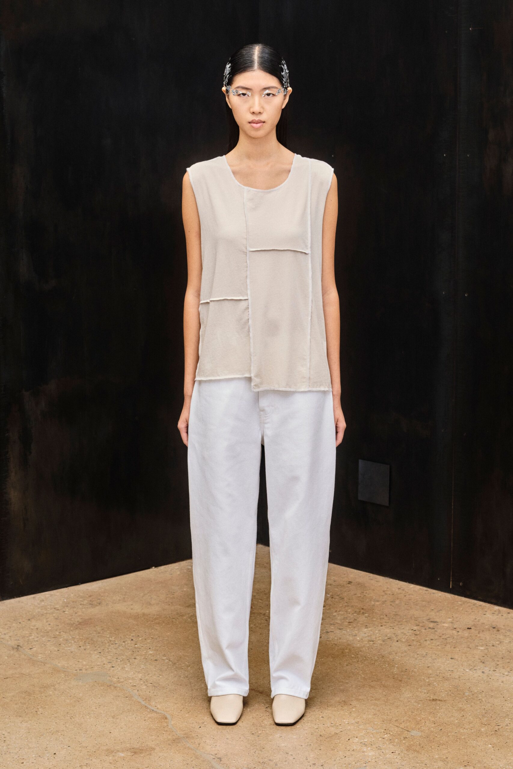 Clear sculpted frames curling into silver clips at the temples. Sheer oyster sleeveless top, layered in offset raw-edged rectangles. Pale straight trousers with a soft washed, almost cloudy finish. Cream square-toe ankle boots. Quiet museum atrium, rain still drying on the concrete outside.