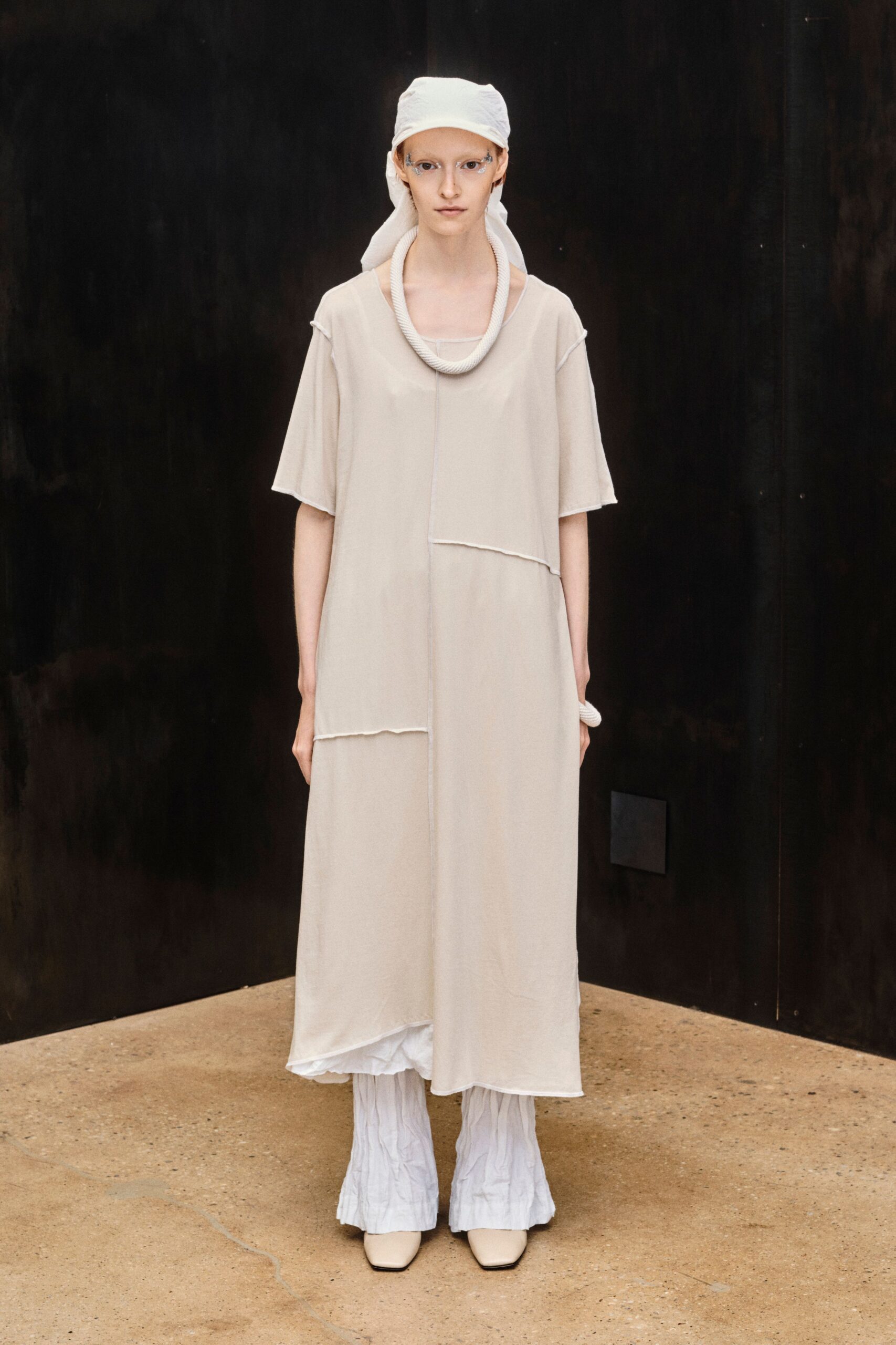 White headscarf tied under a cap, tails falling down the back. Thick ivory rope loop sitting heavy at the collarbone. Milk sheer jersey dress with raw seams and offset patch panels. White crinkled trousers flaring softly below the hem. Cream square-toe boots. Early ferry crossing, salt drying on the rail before breakfast.