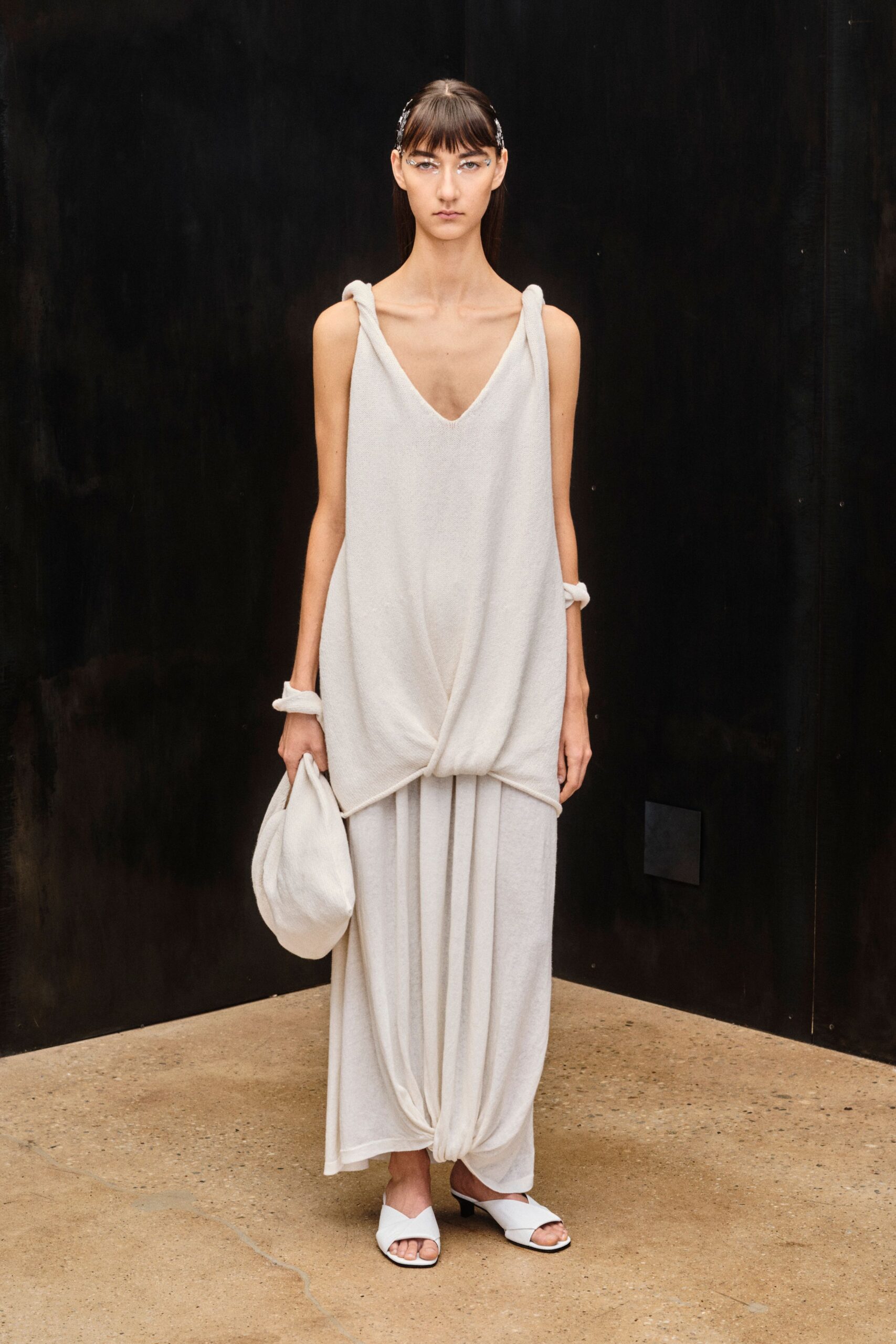 Chalk knit slipdress with knotted rope straps and a low V neck. Soft crescent pouch in the same fuzzy knit. Matching padded cuffs at both wrists. White leather mules with a folded vamp over kitten heels. For crossing a stone courtyard before the spa opens.