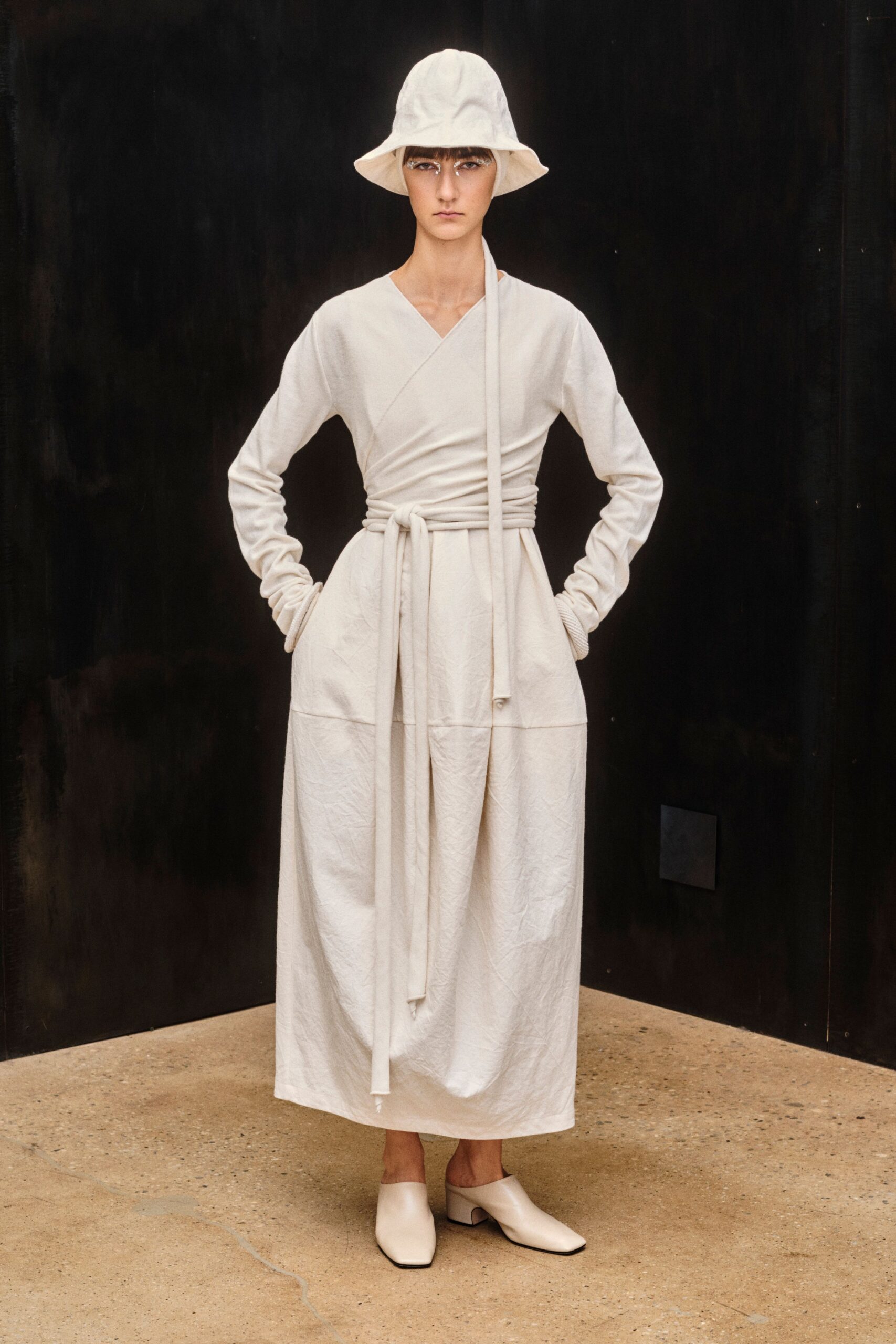 Soft ivory bucket hat with a lightly drooping brim. Chalk wrap dress in crinkled cloth, long ties knotted and dangling past the knee. Cream leather mules with square toes and low block heels. Quiet museum atrium, rain still drying on the stone outside.