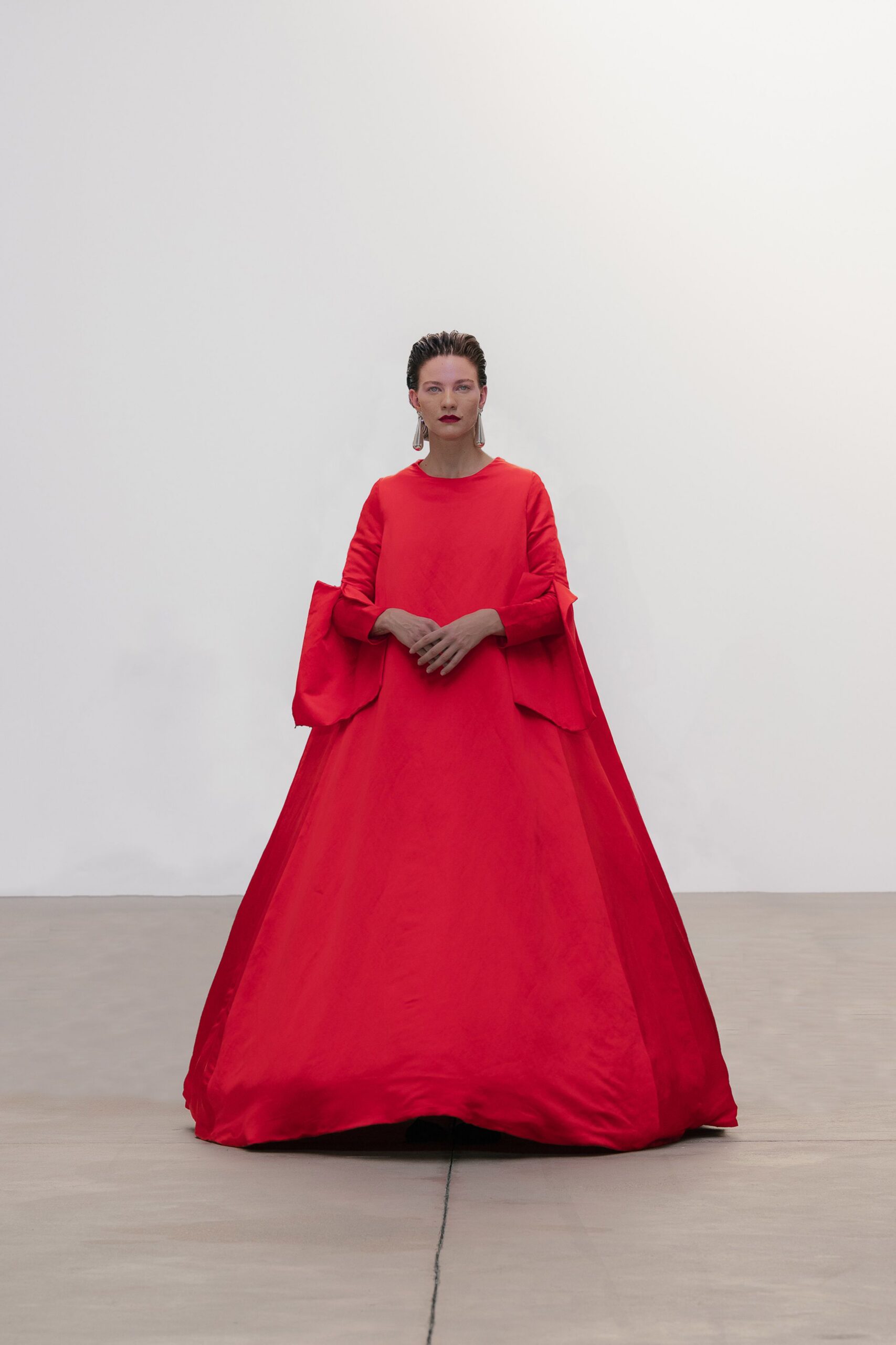 Long silver drop earrings tapering like narrow metal tassels. Vermilion gown in crisp taffeta, balloon skirt and split over-sleeves with raw edges. Crossing a museum atrium after the donor dinner has thinned out.