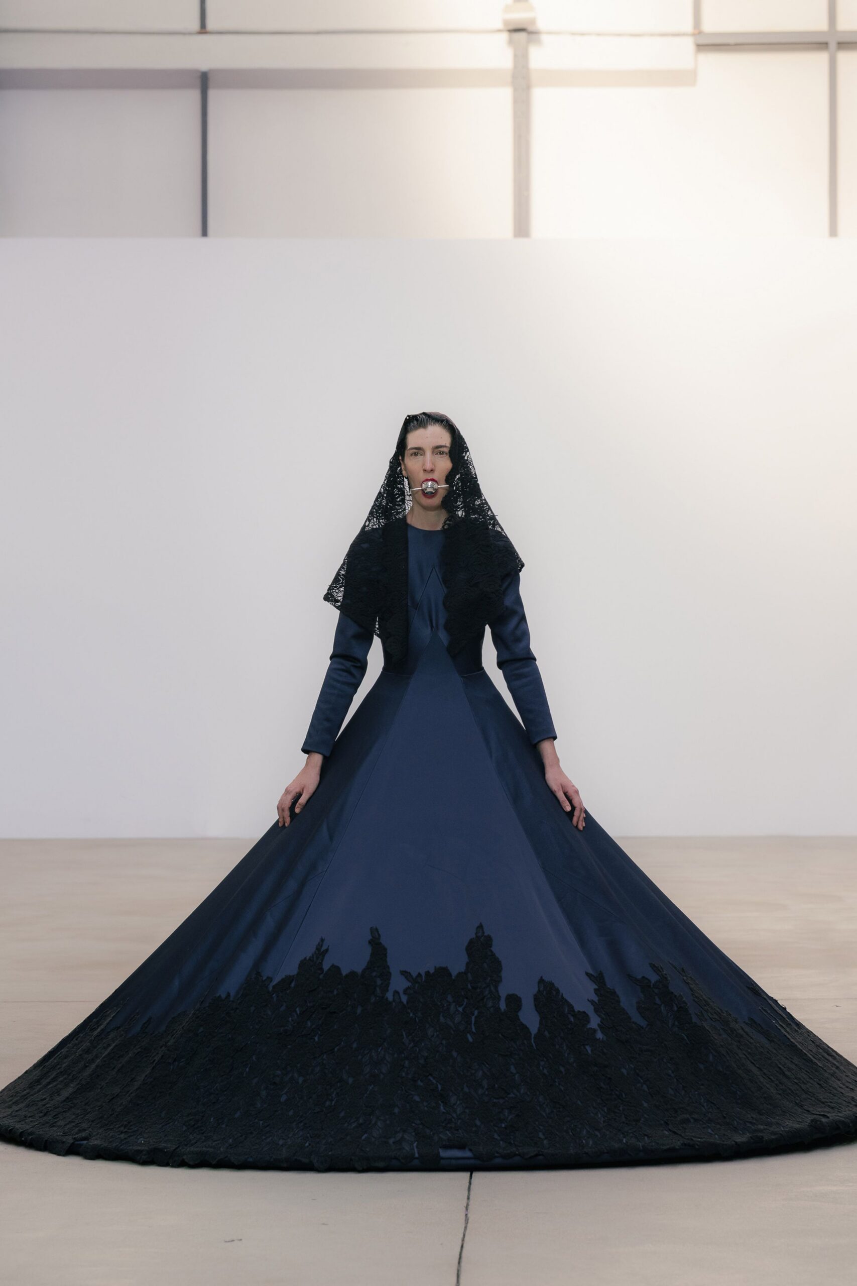 Black lace mantilla falling to the shoulders in a triangular drape. Midnight satin gown, corseted through the waist with a cathedral train. Black floral lace climbing up from the floor in dense silhouette. Silver ball gag held on a narrow strap. Standing in a white chapel after everyone else has gone home.