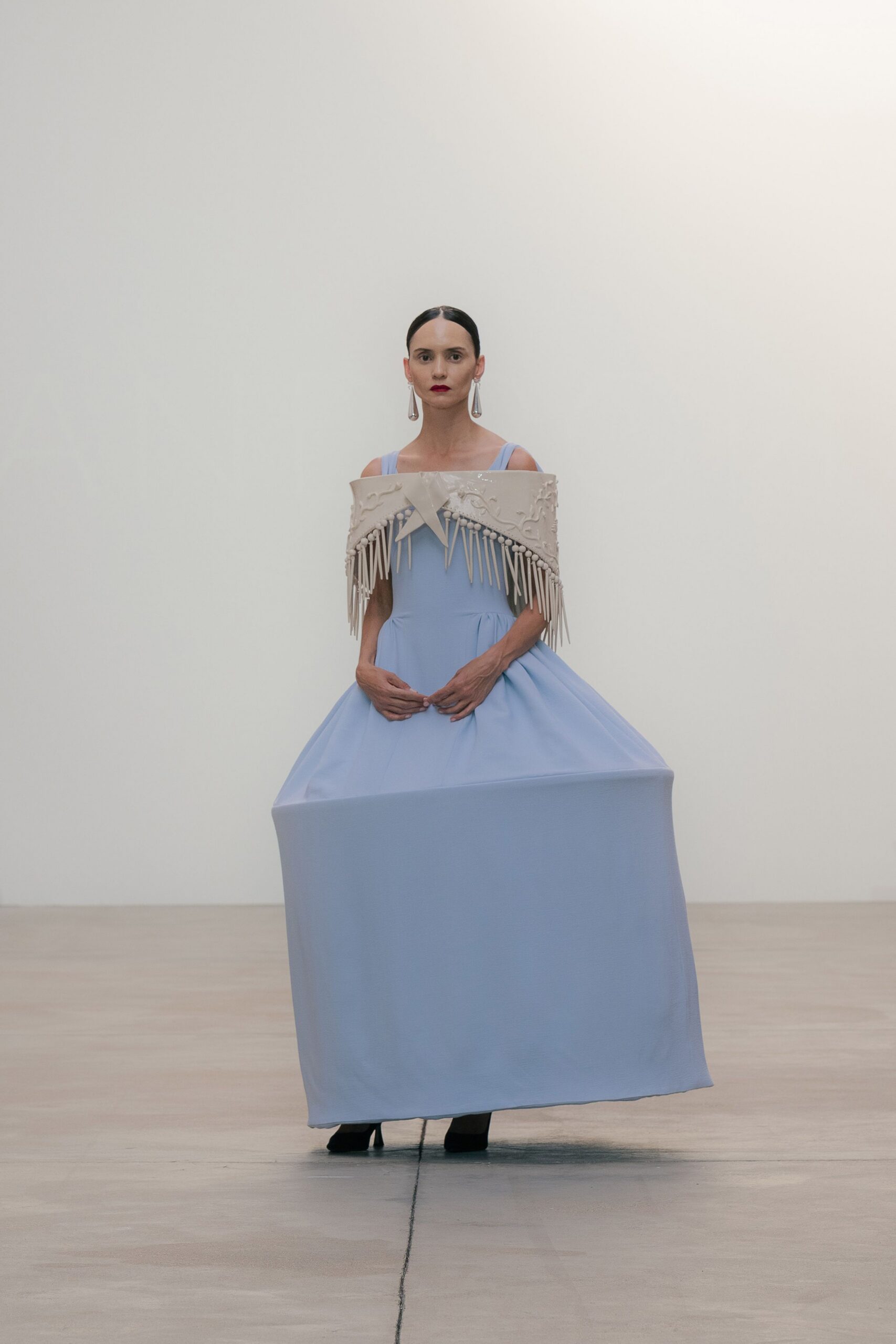 Long teardrop earrings in clear lucite. Glossy ivory sculpted collar with dangling spike fringe and a centered bow. Powder-blue gown cut into a stiff boxy skirt, black shoes just peeking. At the museum after hours, standing very still near the marble plinths.