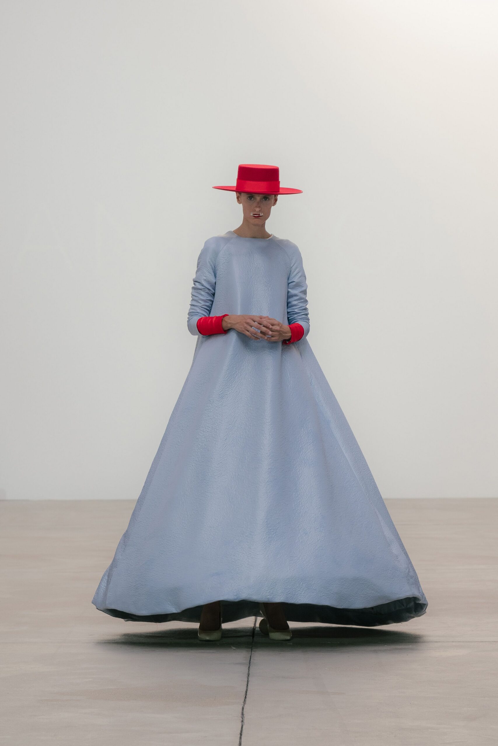 Flat-crowned scarlet hat with a rigid platter brim. Pale blue hammered satin dress ballooning from narrow raglan sleeves. Scarlet cuffs turned back in thick bands at each wrist. Square-toe pumps peeking from the pooled hem. Museum atrium at noon, standing very still while everyone else crosses fast.