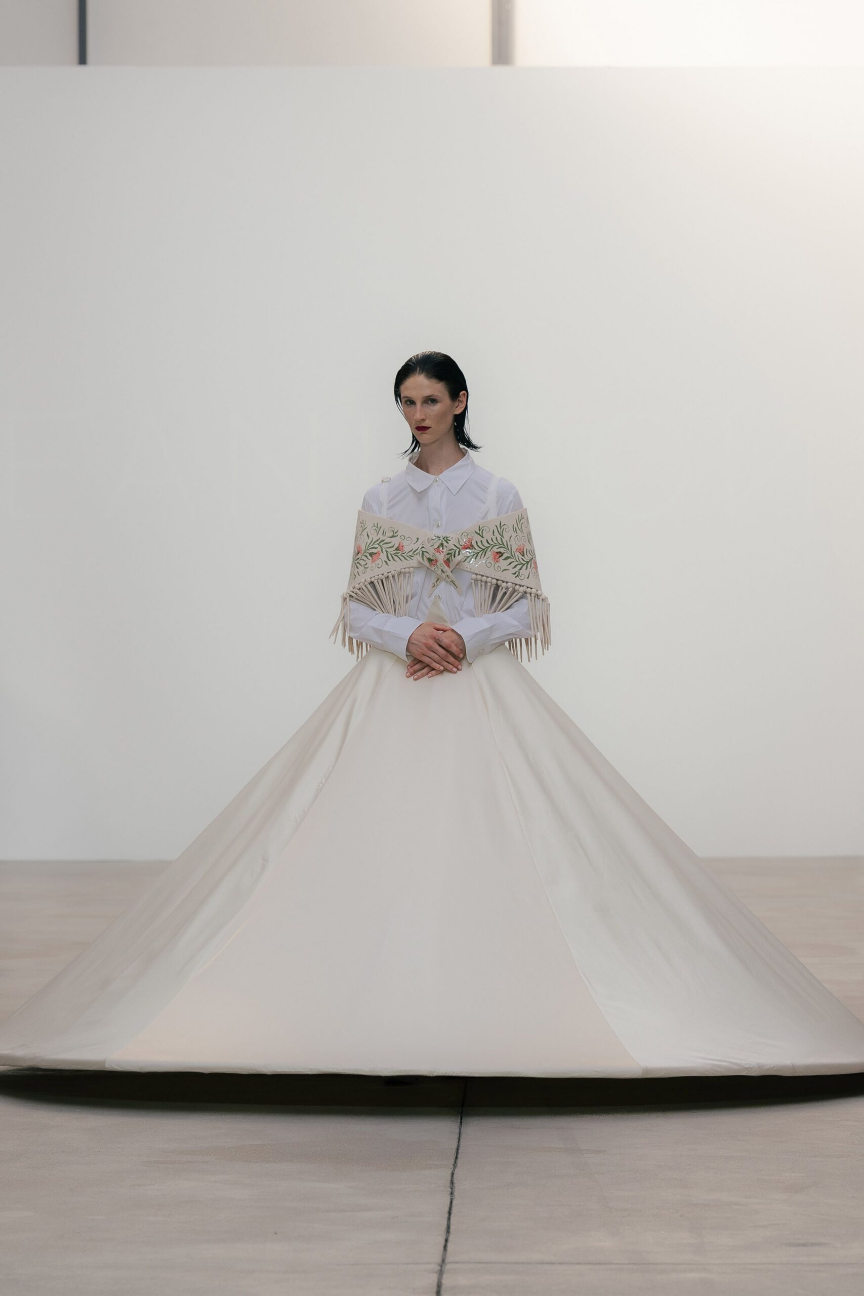 White cotton shirt with a rounded collar and tiny pearl buttons. Cream embroidered shawl slung off the shoulders, knotted fringe hanging straight. Vast ivory skirt in stiff panels, one seam skimming slightly off center. A courthouse wedding before noon, everyone whispering in the marble hall.