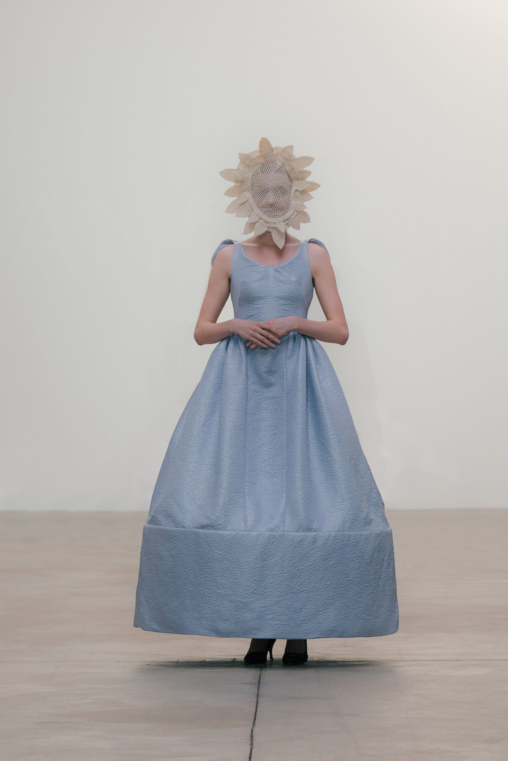 Cream openwork face mask ringed with pointed fabric petals. Pale blue cloqué ball gown, scooped neck and deep turned-up hem. Black pumps barely visible beneath the skirt. Museum atrium hush, standing still while the florist finishes the installation.