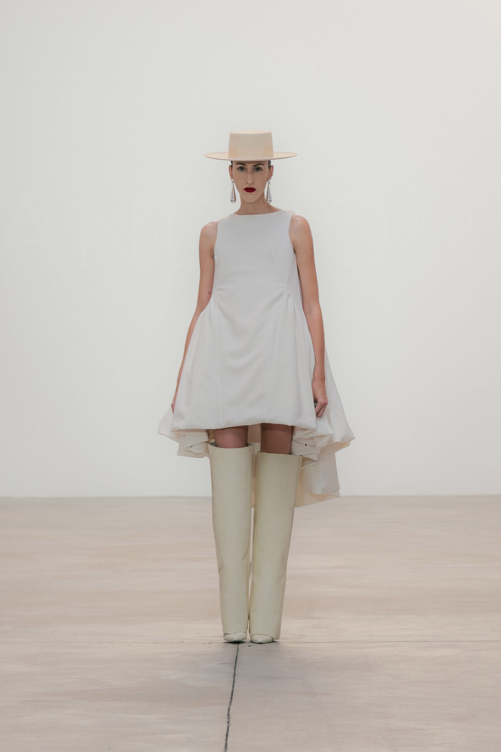 Flat-crowned ivory hat with a razor-wide brim. Long silver teardrops swinging past the jaw. White trapeze mini dress, side panels falling into a trailing back hem. Ivory knee-high boots with blunt square toes. Crossing a chapel courtyard after the ceremony, lipstick still exact.