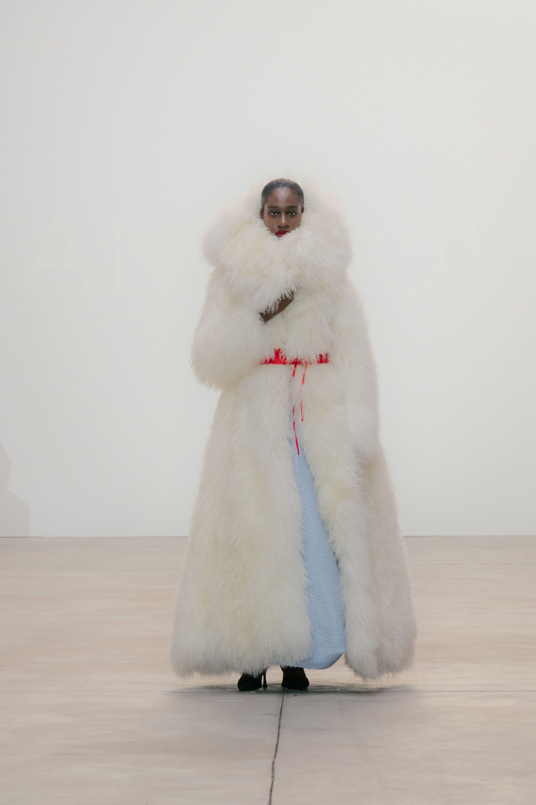 Floor-length ivory shag coat with a huge clouded collar swallowing the jaw. Thin scarlet cord belt, ends hanging loose at the front. Powder-blue textured column dress peeking through the opening. Black pointed boots barely visible under the hem. Crossing a frozen hotel lobby still wrapped in the duvet.