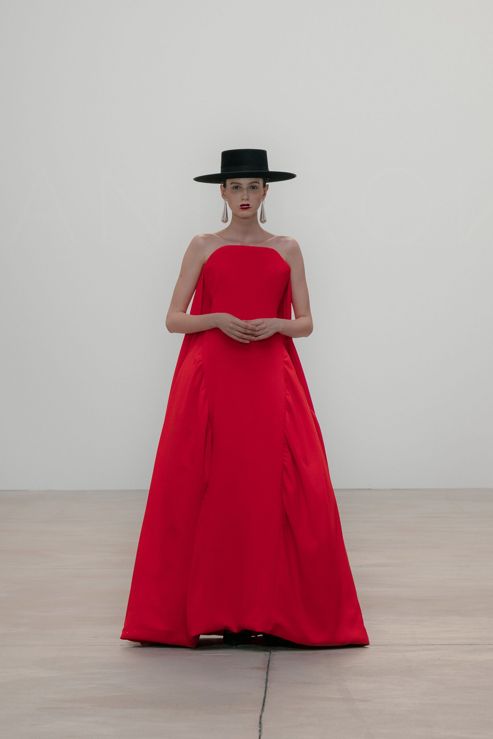 Flat-crowned black hat with a rigid dinner-plate brim. Long silver teardrops grazing the jawline. Scarlet strapless column gown with cape panels falling from the side seams. Black toes barely visible under the pooled hem. For crossing a museum atrium after everyone has already turned to look.