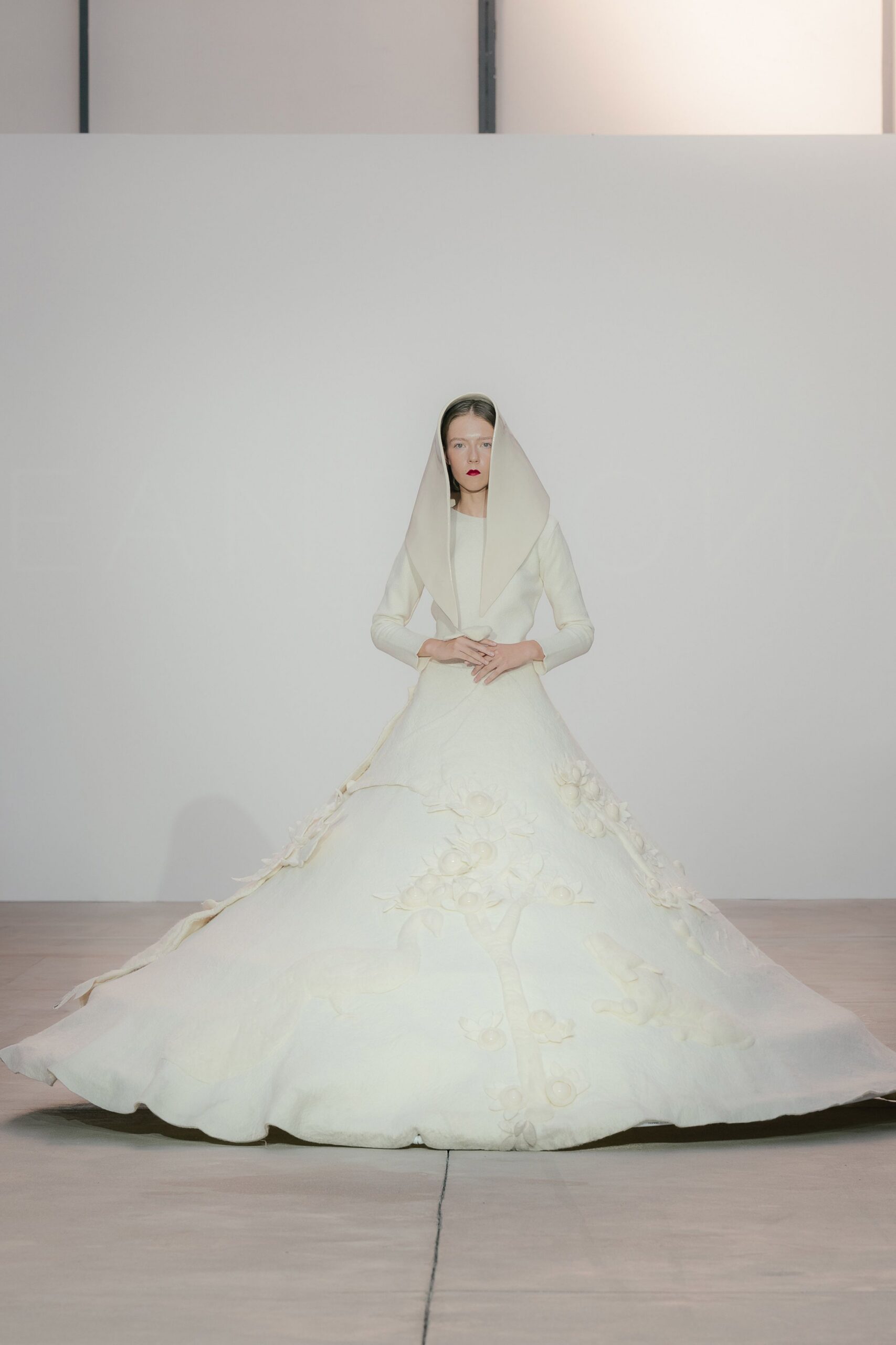 Ivory felt hood falling in two long pointed panels. Sculpted ivory gown with a nipped waist and three-quarter sleeves. Bell-shaped skirt in dense wool, strewn with raised floral appliques. In the chapel after everyone else has gone to the reception.
