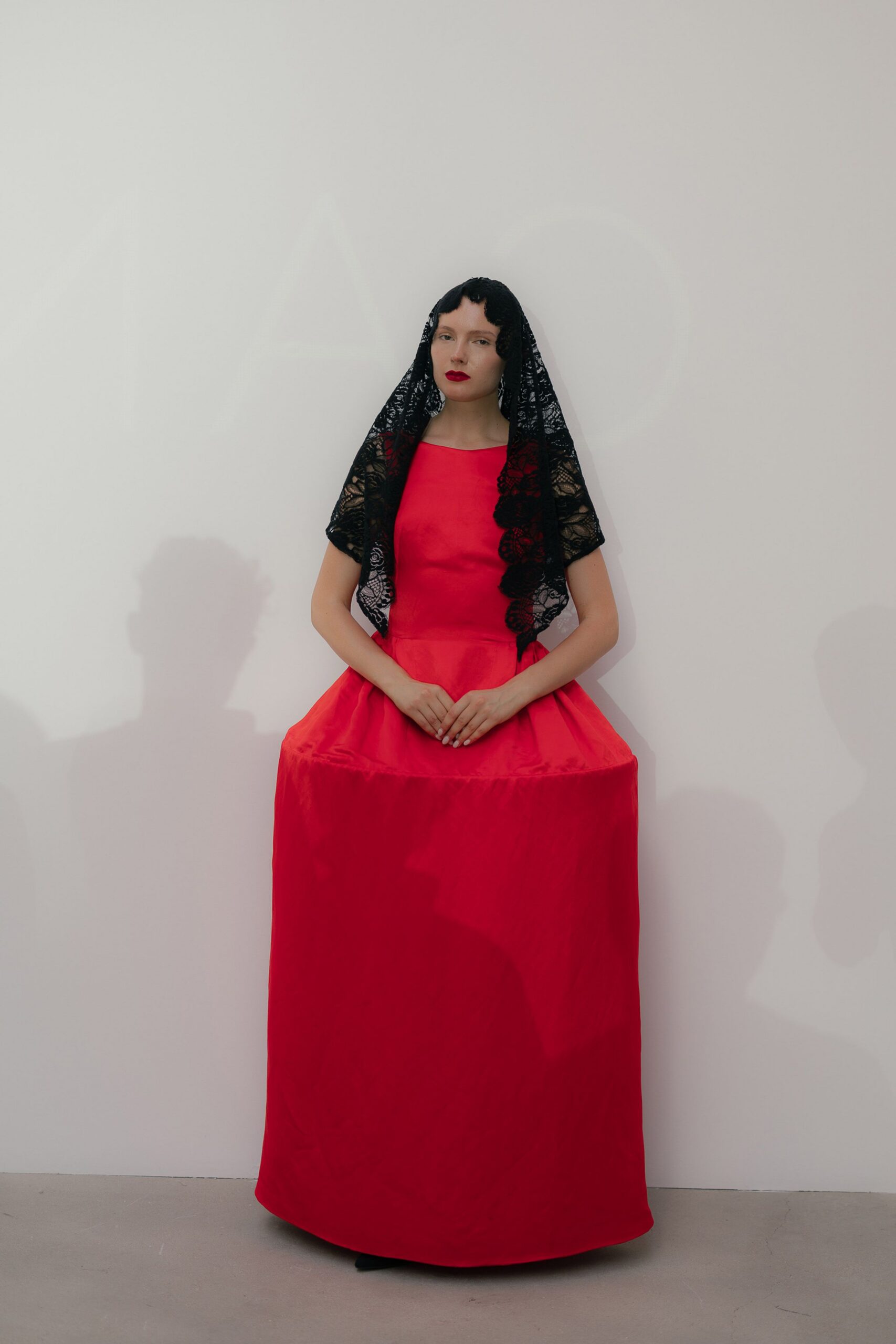 Black floral lace mantilla, scalloped circles running down one side. Cherry duchesse satin column gown with a stiff panniered hip shelf. Black pointed toes barely peeking below the hem. Museum chapel hush, right before the tour group turns the corner.
