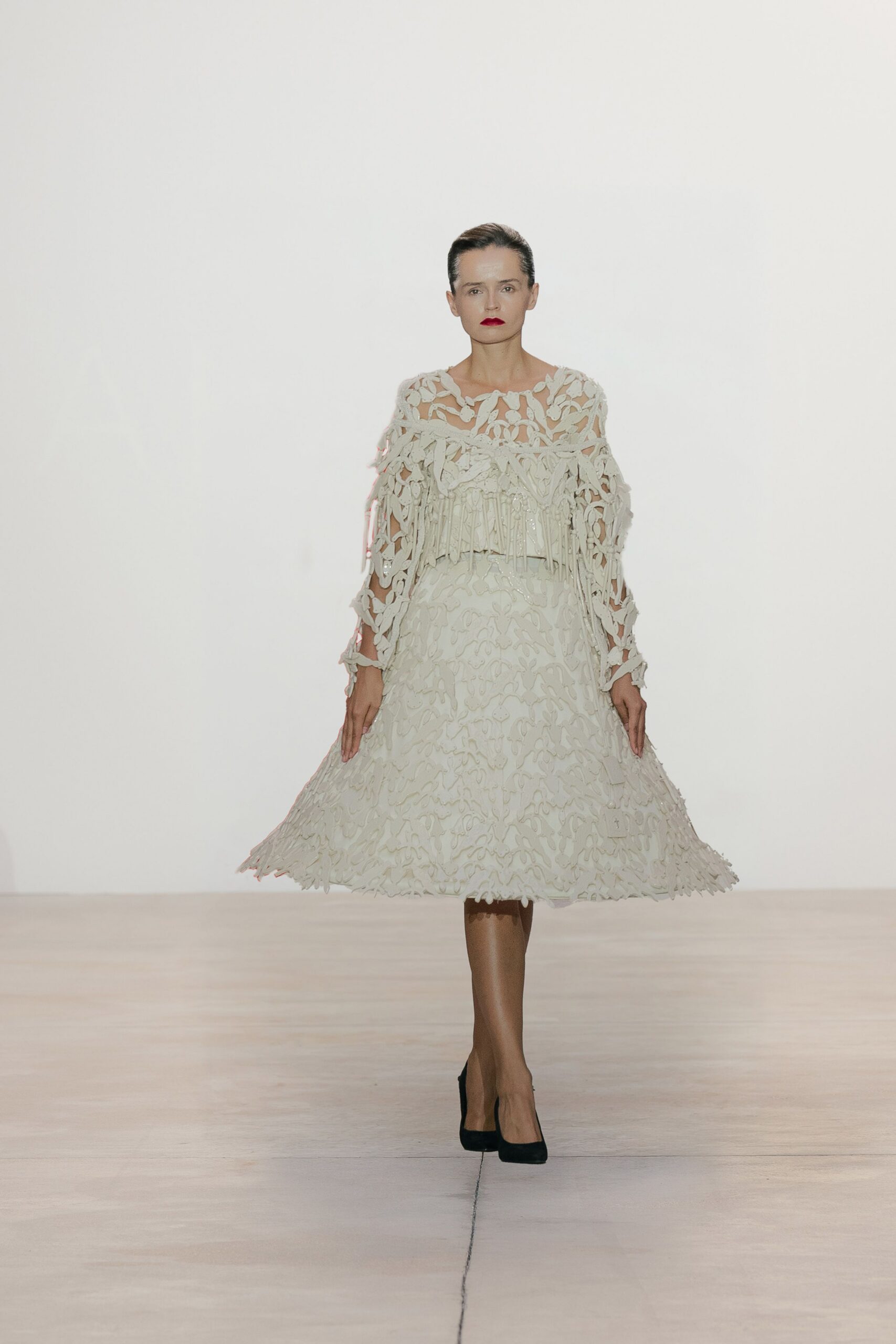 Ivory cutwork dress with looping appliqué and a fringe band across the bust. Black suede pumps under sheer brown tights. Tiny ivory box pouch hanging at the right hip. Church hall wedding between services, lipstick still too bright for daylight.