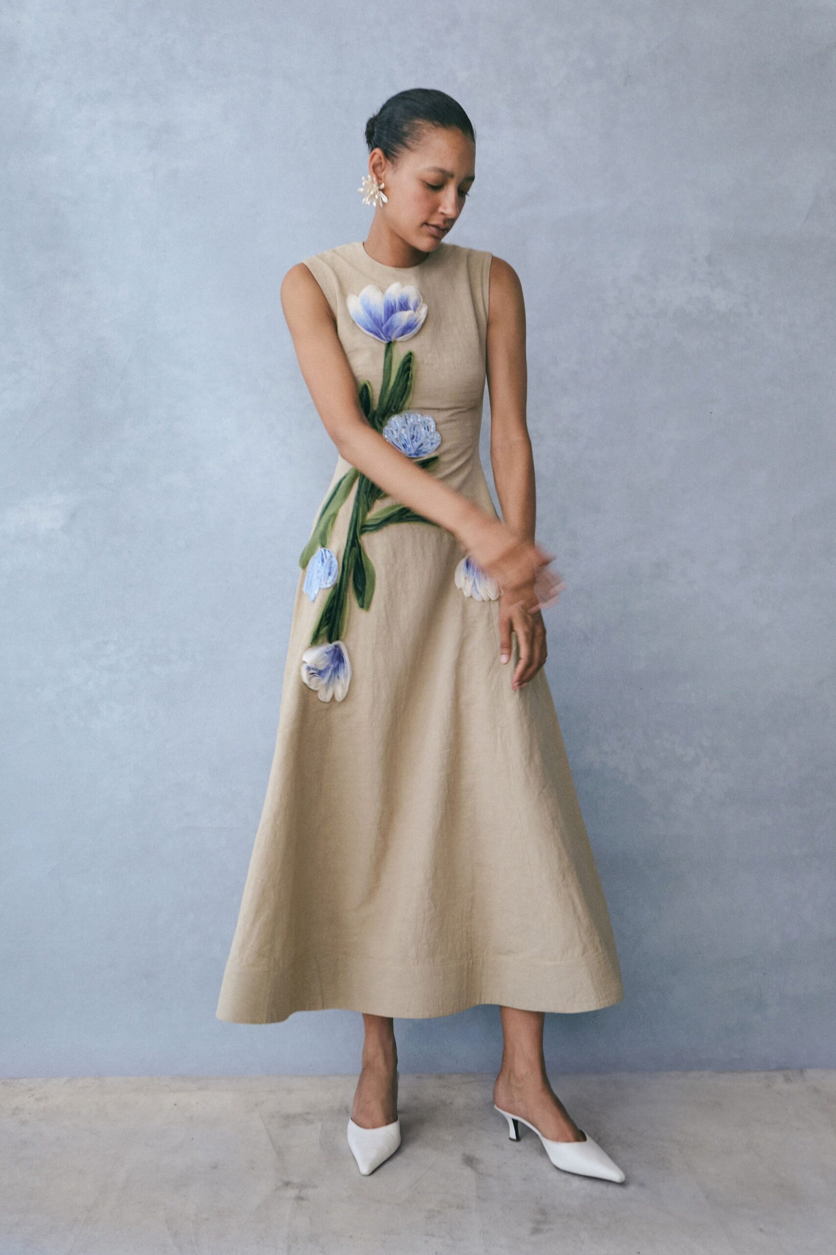 Oat linen sleeveless dress with oversized blue floral appliques trailing downward. Milky petal cluster earrings. White pointed mules on a slim kitten heel. Afternoon wedding in a conservatory, someone has just opened the side doors.