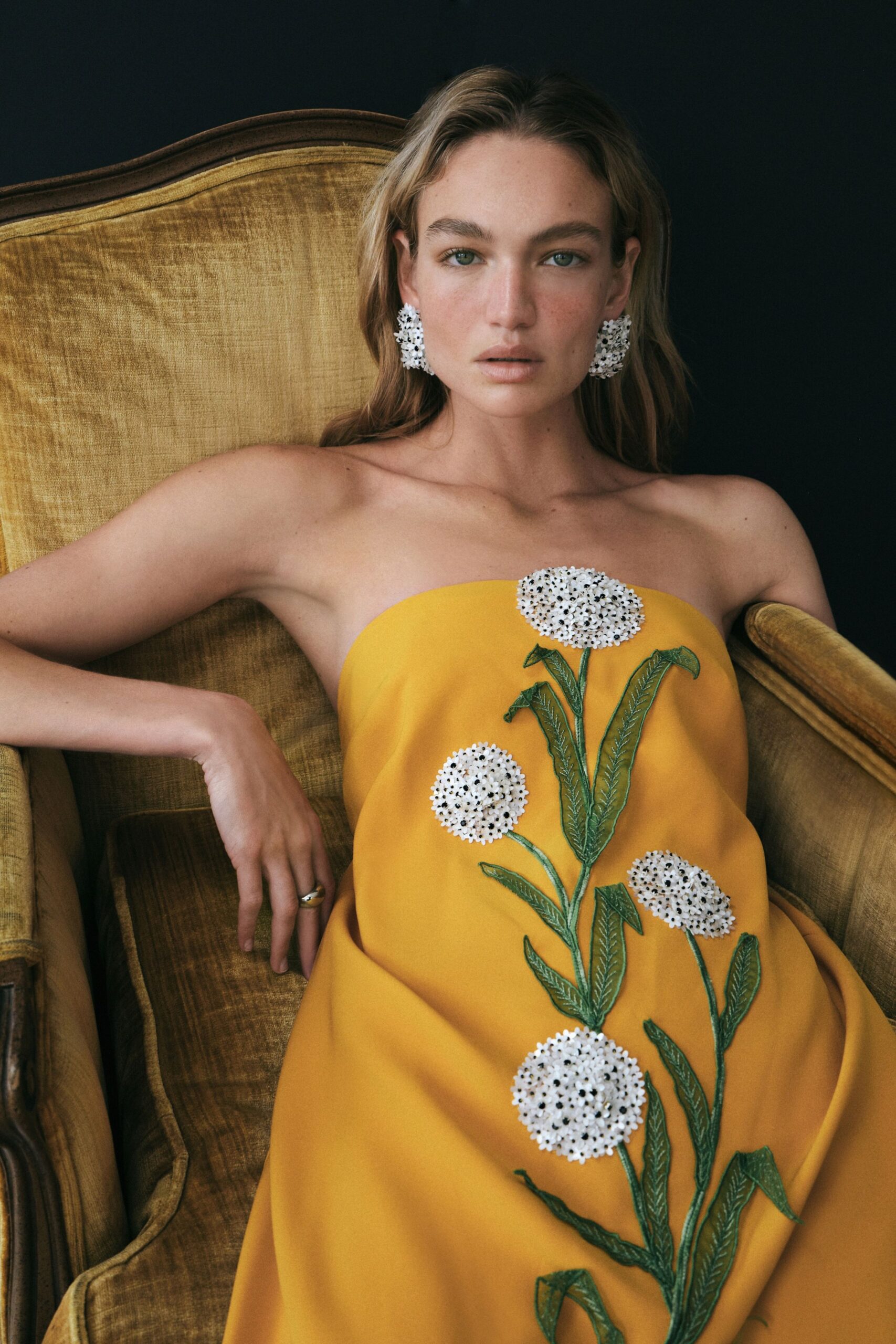 White clustered floral earrings with peppered black bead centers. Marigold column gown, oversized white flower appliques climbing on stitched green stems. Thick gold ring on the slack middle finger. Sunroom portrait after lunch, velvet chair nap lines still on your arm.
