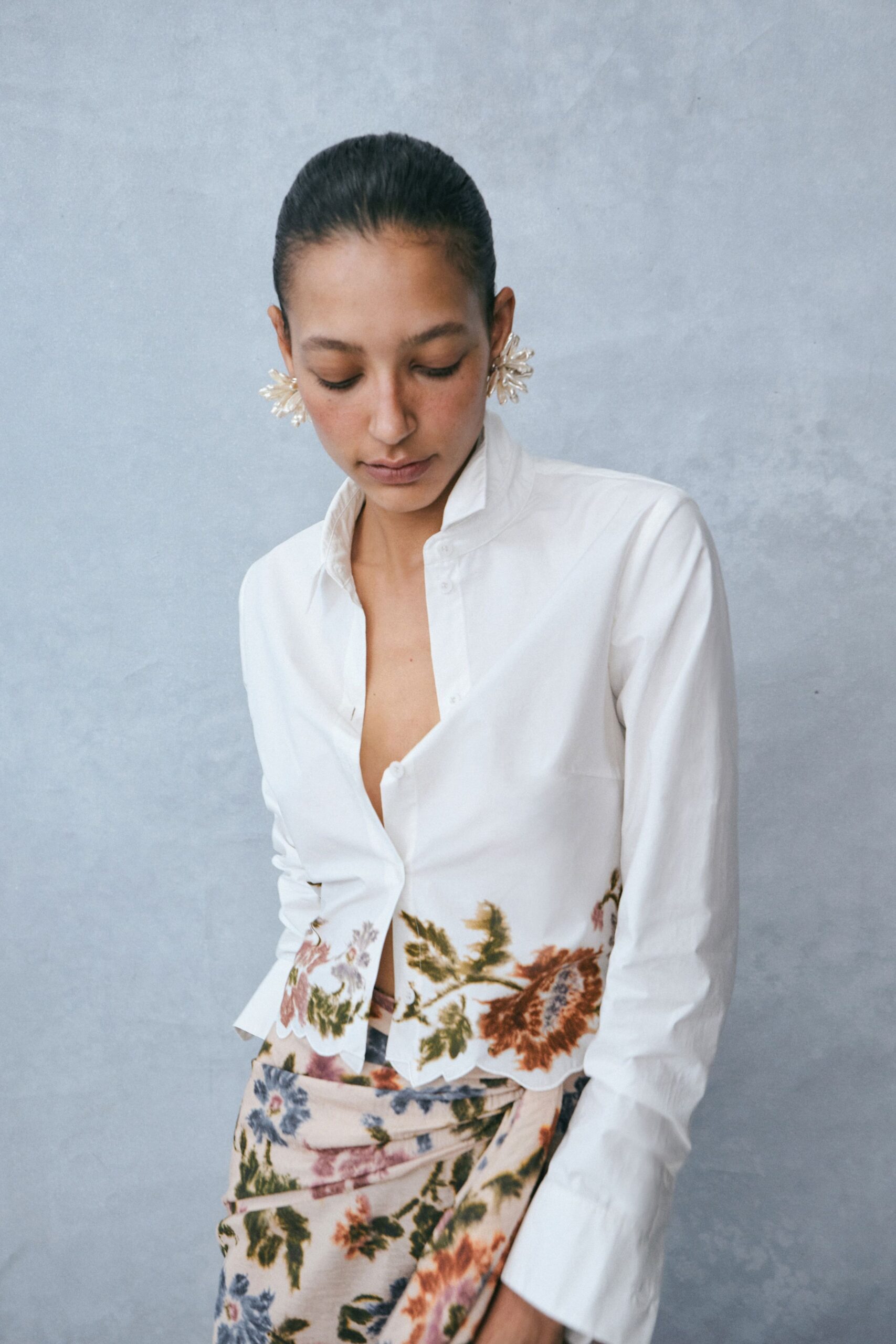 Milky petal-cluster earrings flaring just past the earlobes. White cotton shirt with a popped collar and scalloped floral hem. Blush wrap skirt in faded tapestry florals, folded tight at the hip. Sunday lunch on a porch where the tablecloth keeps lifting.