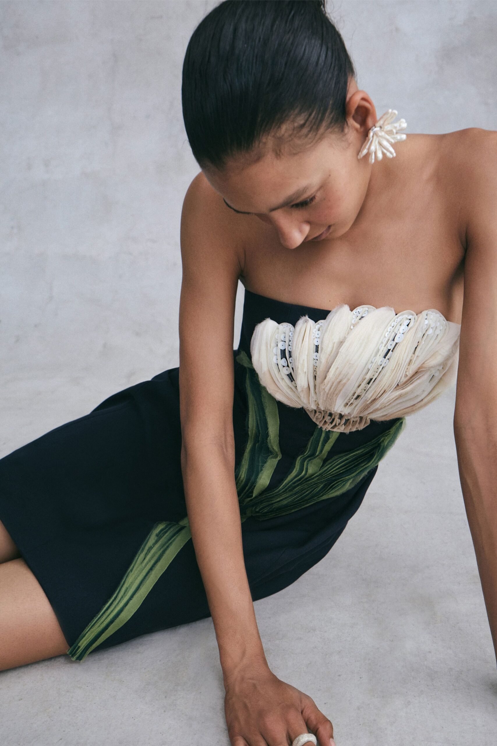 Pearl-cluster earrings fanning like small white petals. Black column dress with a shell-pink pleated bust, sequins tucked between the folds. Chunky white ring at the bent hand. Afternoon wedding in a concrete house, leaning down to pick up a dropped place card.