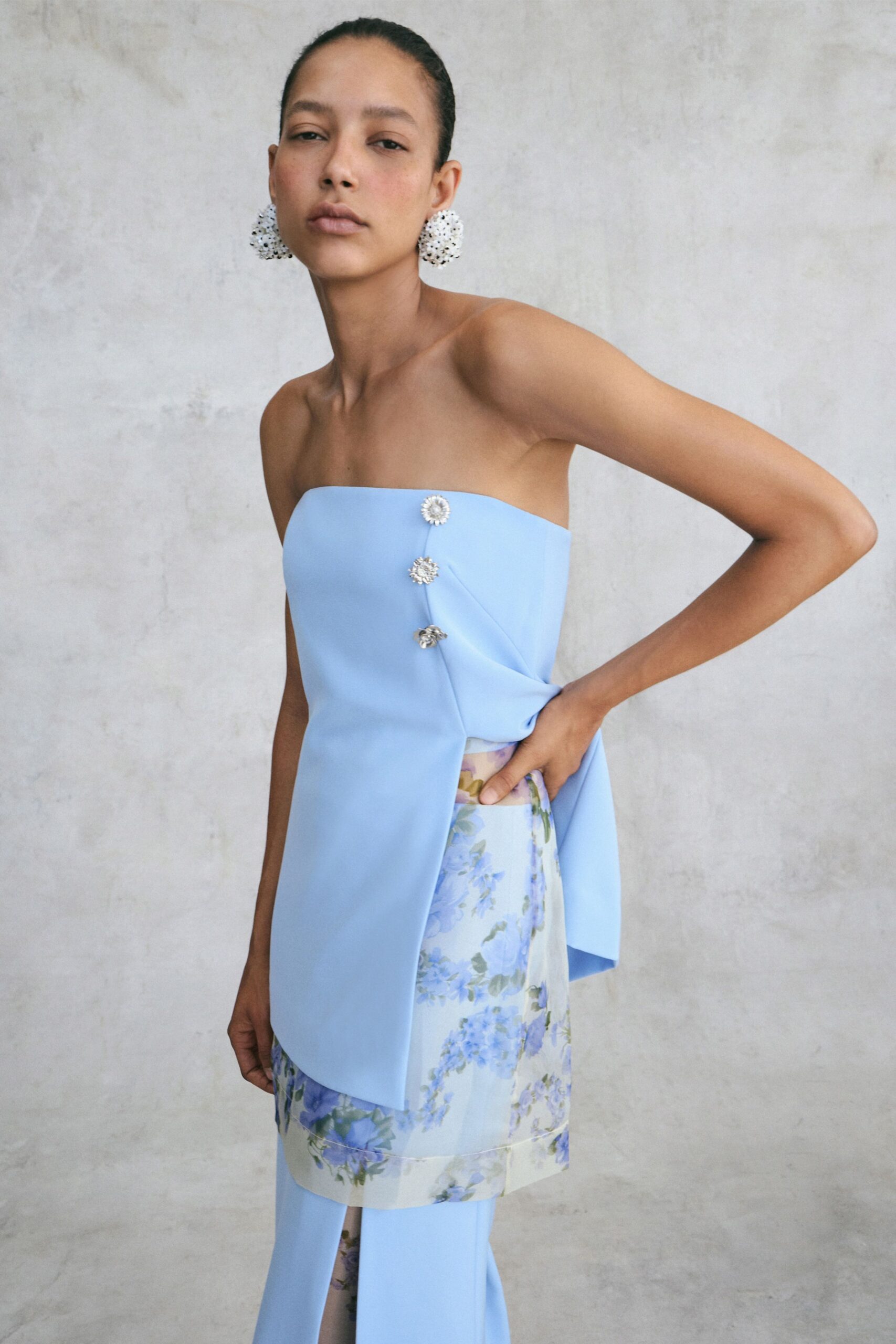 White clustered drop earrings with a beaded, sea-anemone surface. Powder-blue bonded panel wrapping the torso, pinned with three crystal rosettes. Pale floral skirt peeking through the side slit, glossy at the hem. Garden wedding noon, standing where the shade ends on the terrace.