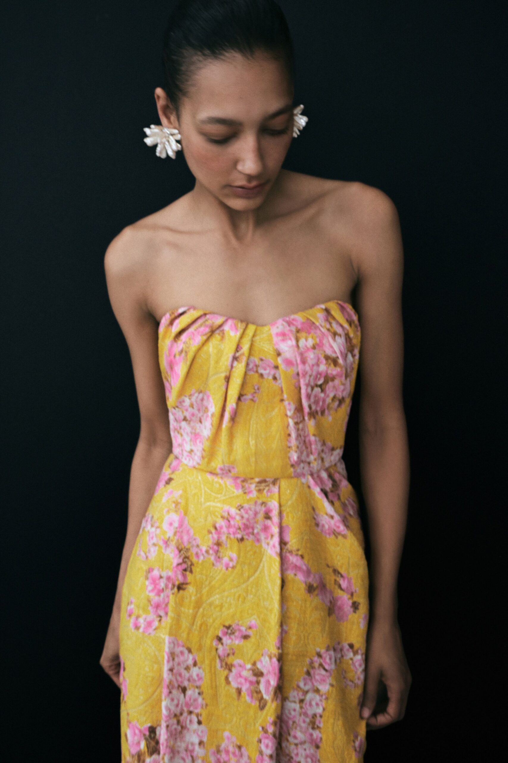 White petal-cluster earrings, slightly oversized against the jaw. Strapless marigold jacquard dress with pink blossom sprays and soft bust pleats. Garden wedding between rain showers, shoes sinking a little into the grass.