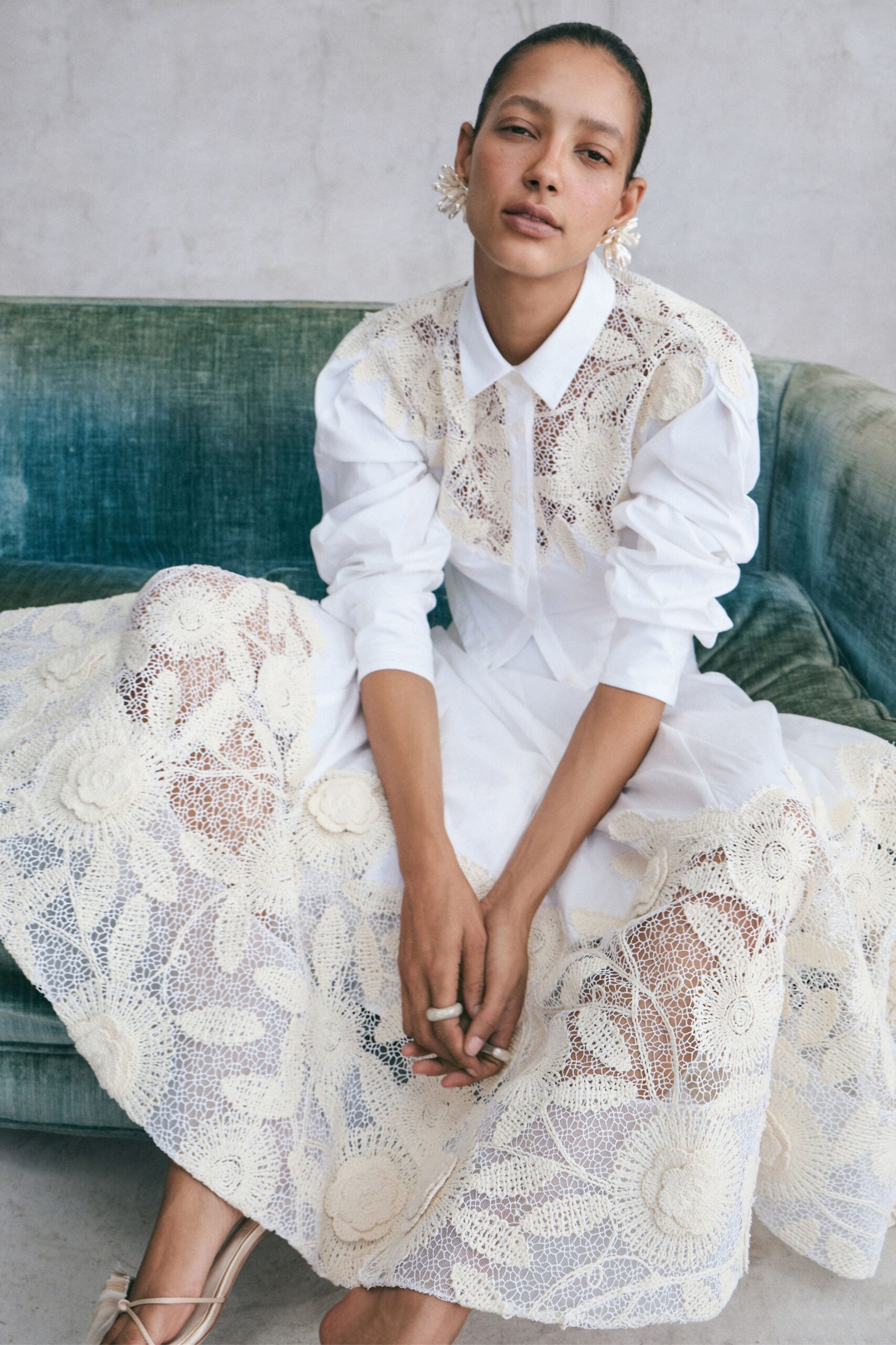 White poplin shirt with a pointed collar and gathered sleeves. Full ivory lace skirt pieced from oversized floral crochet medallions. Barely there thong sandals in pale leather. Milky clustered drop earrings. Sunday hotel lounge, waiting for the tea tray to arrive.