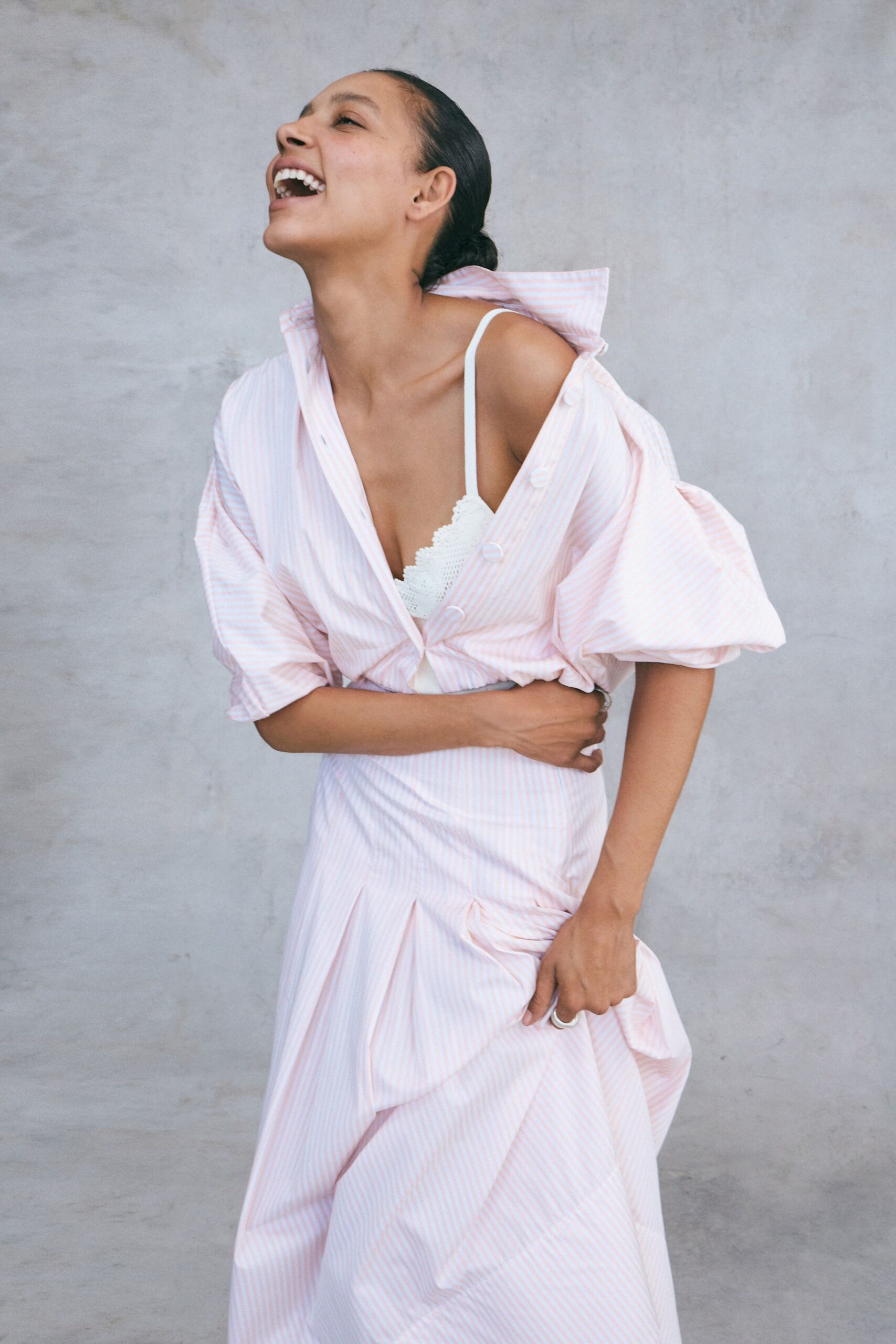 Blush-and-white striped shirtdress with a popped collar and puffed elbow sleeves. White crochet bralette peeking through the open placket. Plain silver band on the gripping hand. Laughing on a terrace after lunch, still holding the skirt out of the breeze.