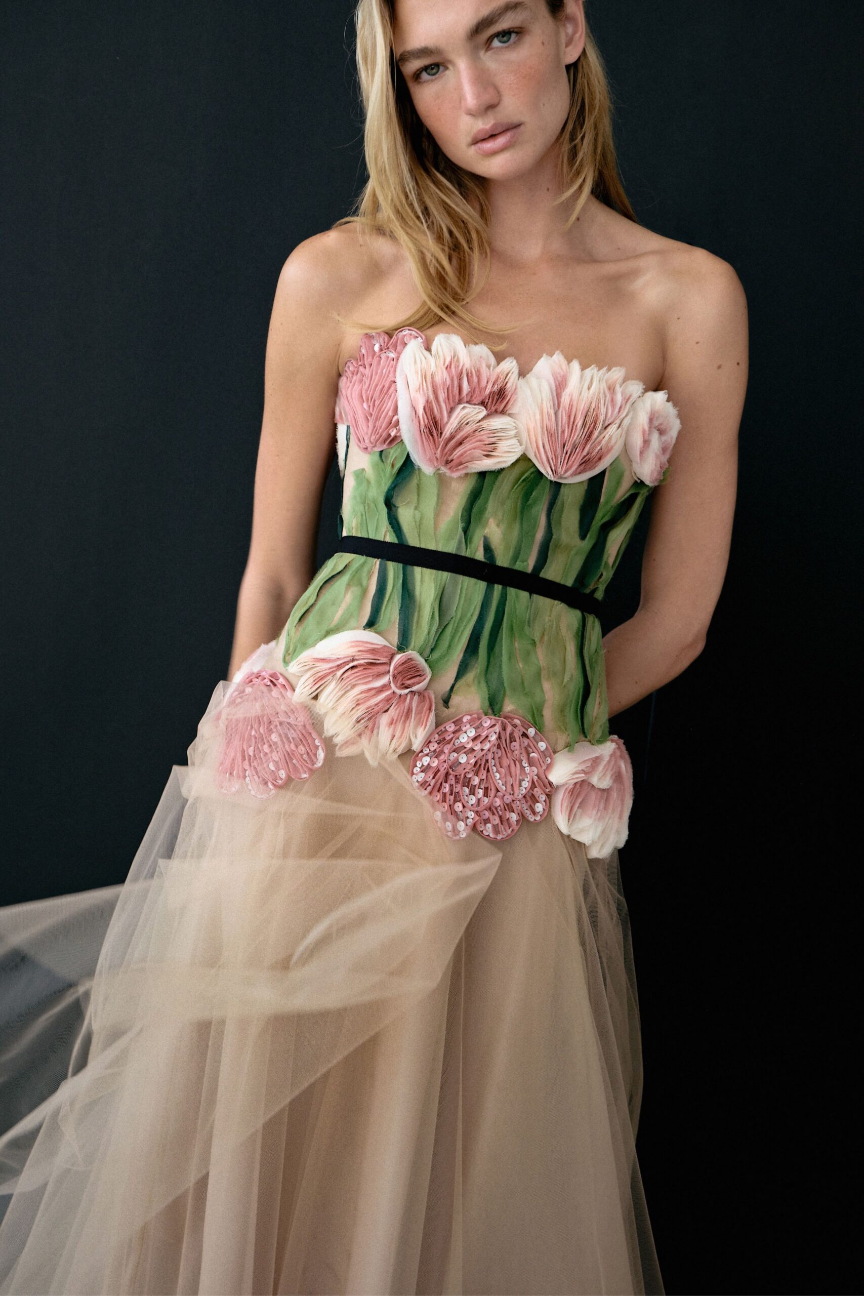 Strapless bodice built from pink petal appliqué over painted green stems, black band at the waist. Nude tulle skirt falling in cloudy layers, one sheer panel lifting away at the hip. For standing in a glasshouse after closing, petals swept into the drains.