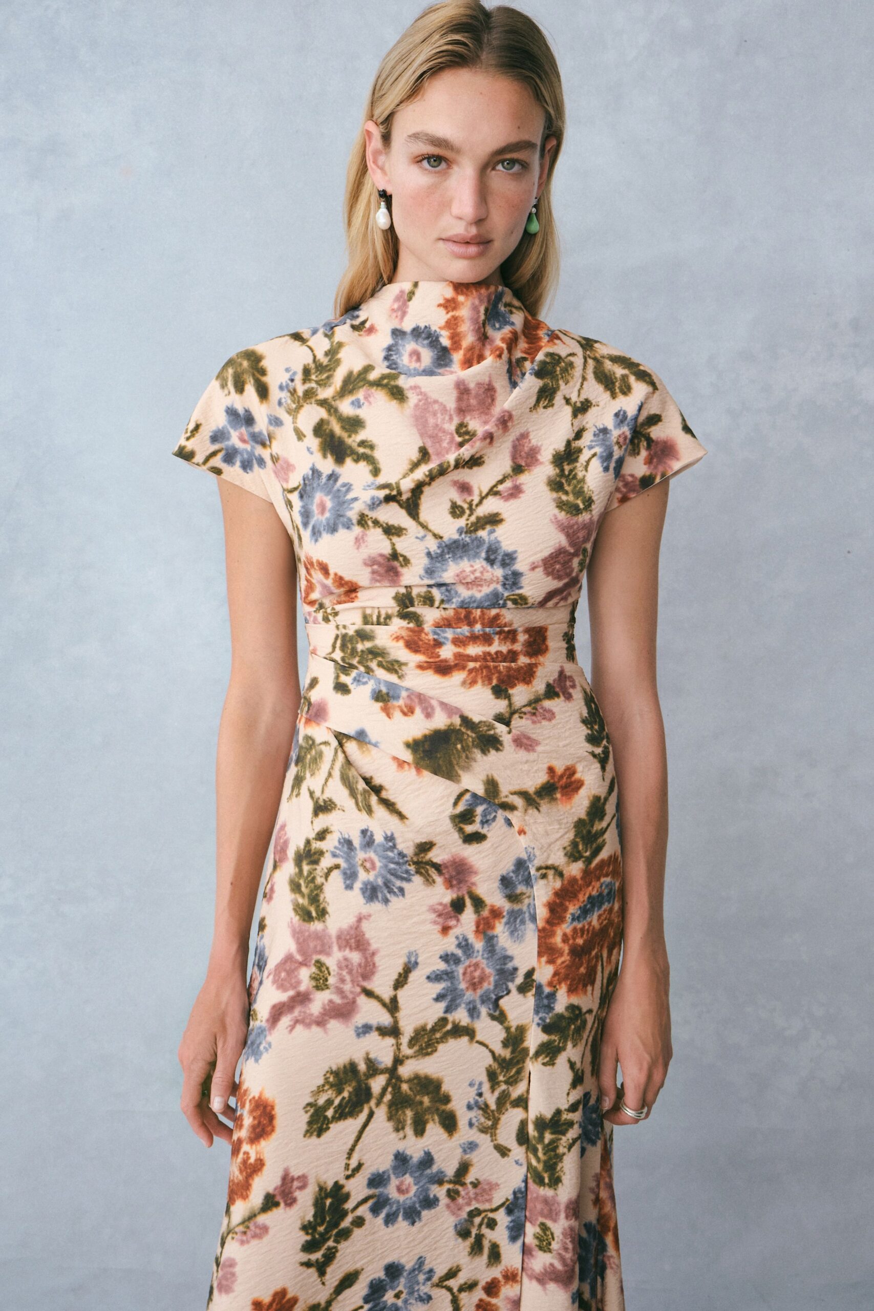 Mismatched drop earrings, one pearl and one green teardrop bead. Cream crinkled dress scattered with dusty blue and rust florals, cap sleeves and a wrapped high neck. Bias-cut bands crossing the waist, one seam landing slightly off center. Slim silver ring. Garden wedding before noon, grass still damp through the soles.