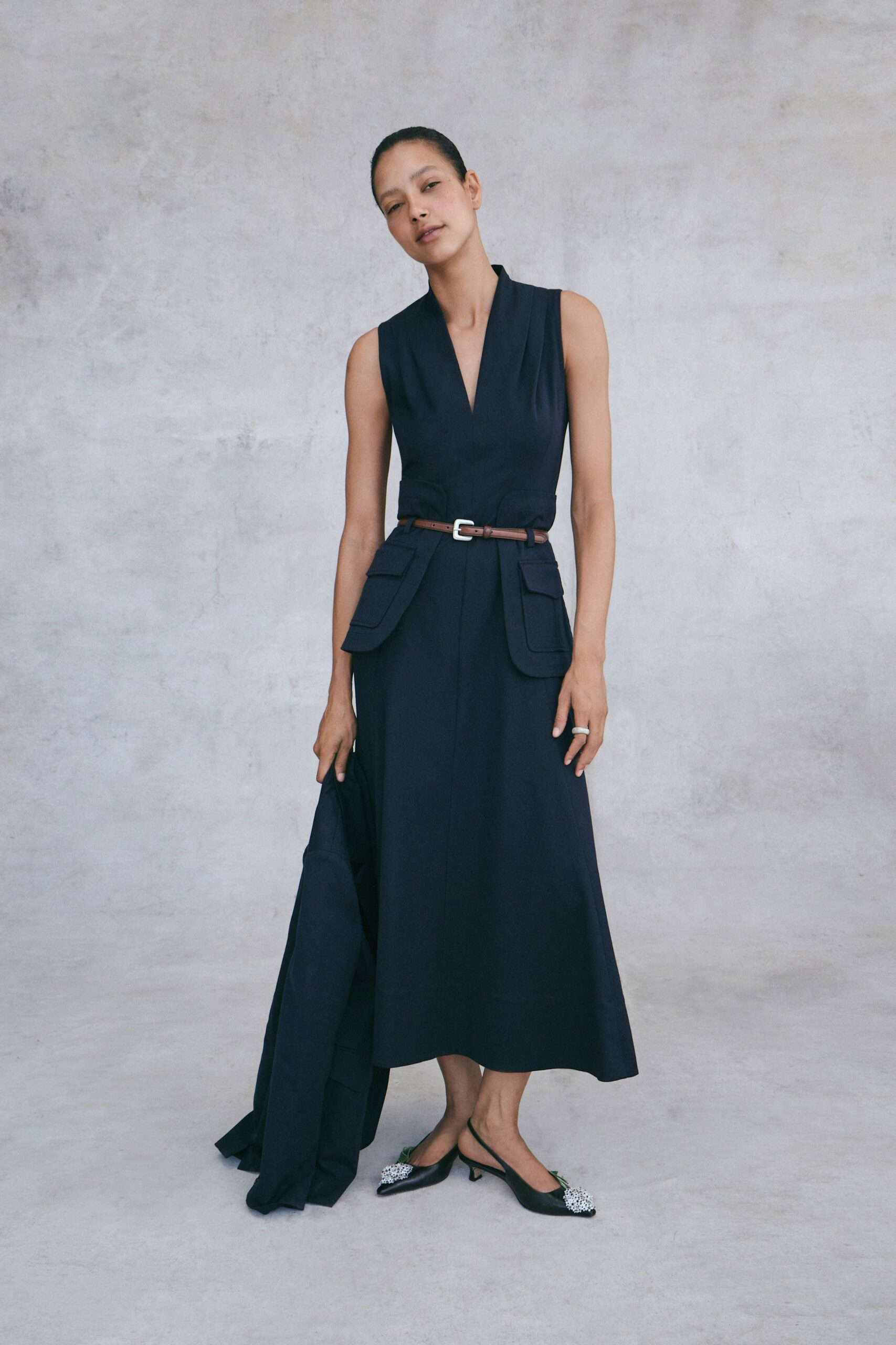 Inky sleeveless dress with a plunging shawl collar and flap hip pockets. Narrow brown leather belt with a square silver buckle. Matching dark coat carried loose in one hand. Black slingbacks with crystal-cluster toes and kitten heels. For arriving early to a courtyard wedding before the chairs are set.